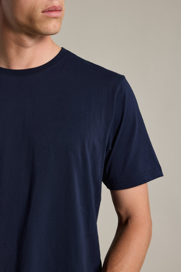 A man models the Barkers Organic Crew Tee in dark blue, made from organic cotton. Shown from shoulders to mid-arm and facing left against a neutral background, his face is partly visible and he doesn’t look at the camera.