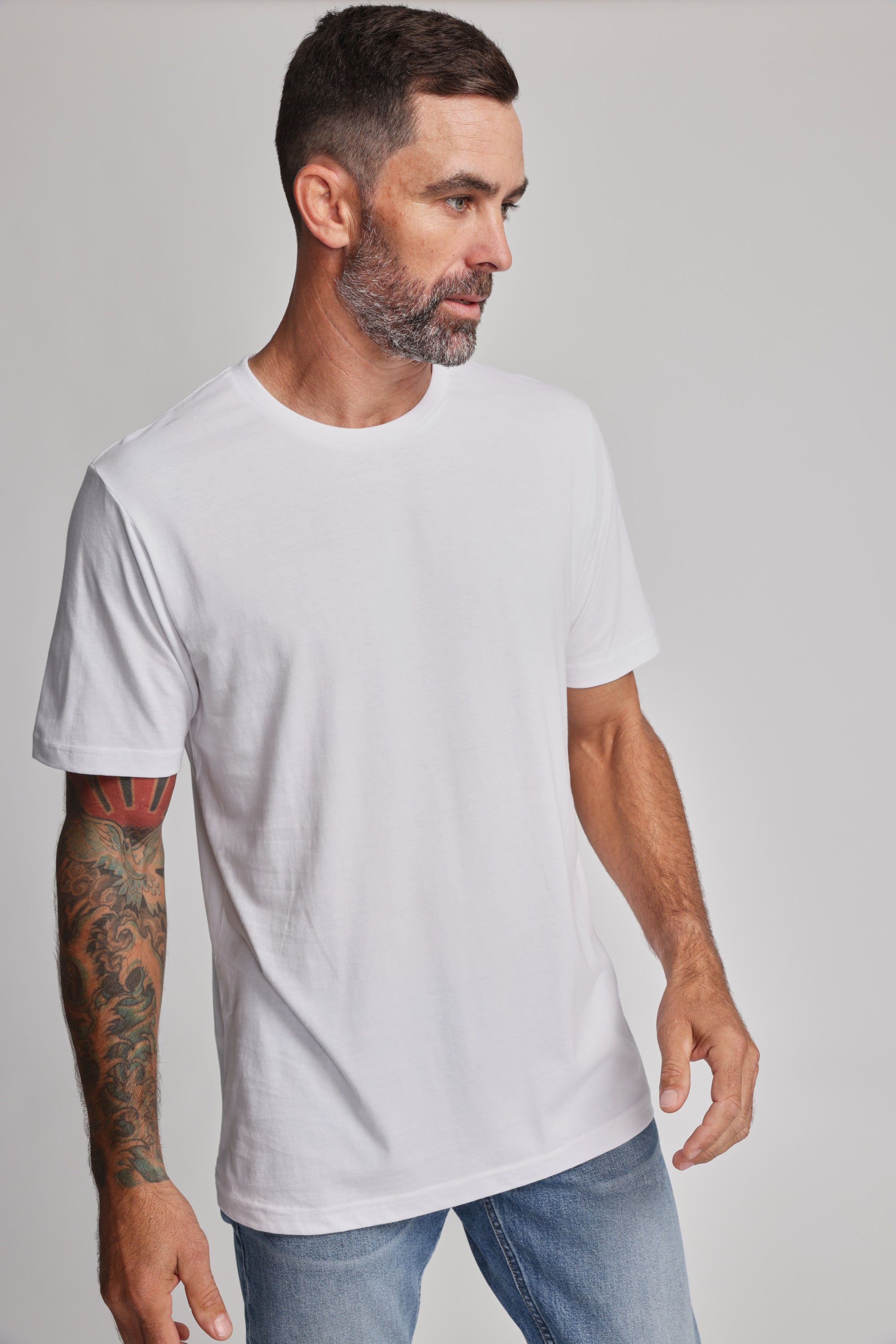 A man with short dark hair and a beard models the Barkers Organic Crew Tee in white with light blue jeans. He displays a colorful tattoo sleeve while standing against a plain light background, gazing to his left.