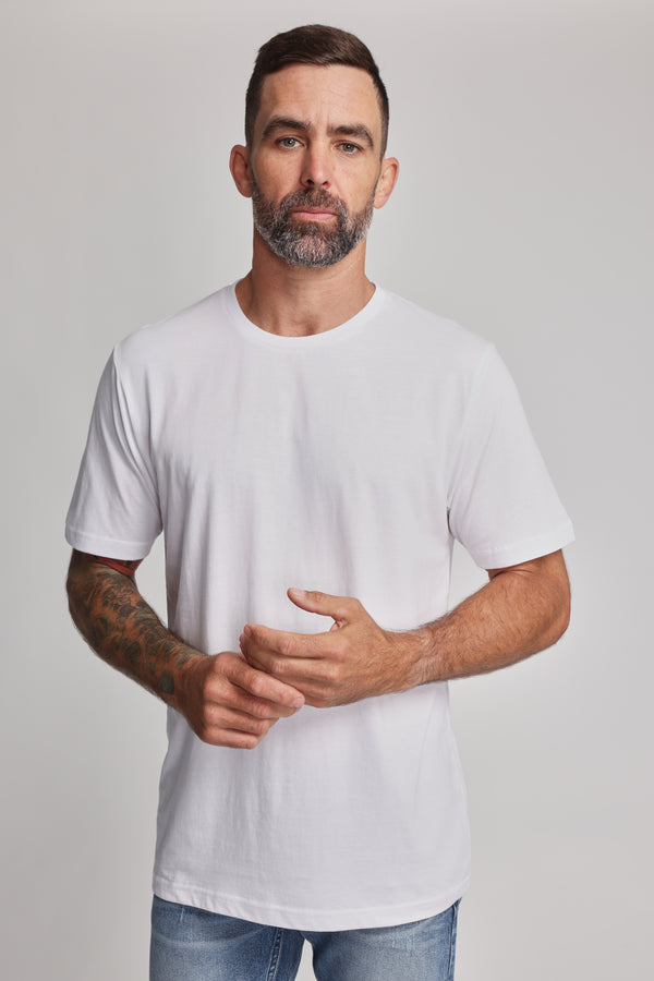 A man with short dark hair, a beard, and a tattoo sleeve stands against a plain background, wearing the Barkers Organic Crew Tee and blue jeans. He faces the camera with a neutral expression.