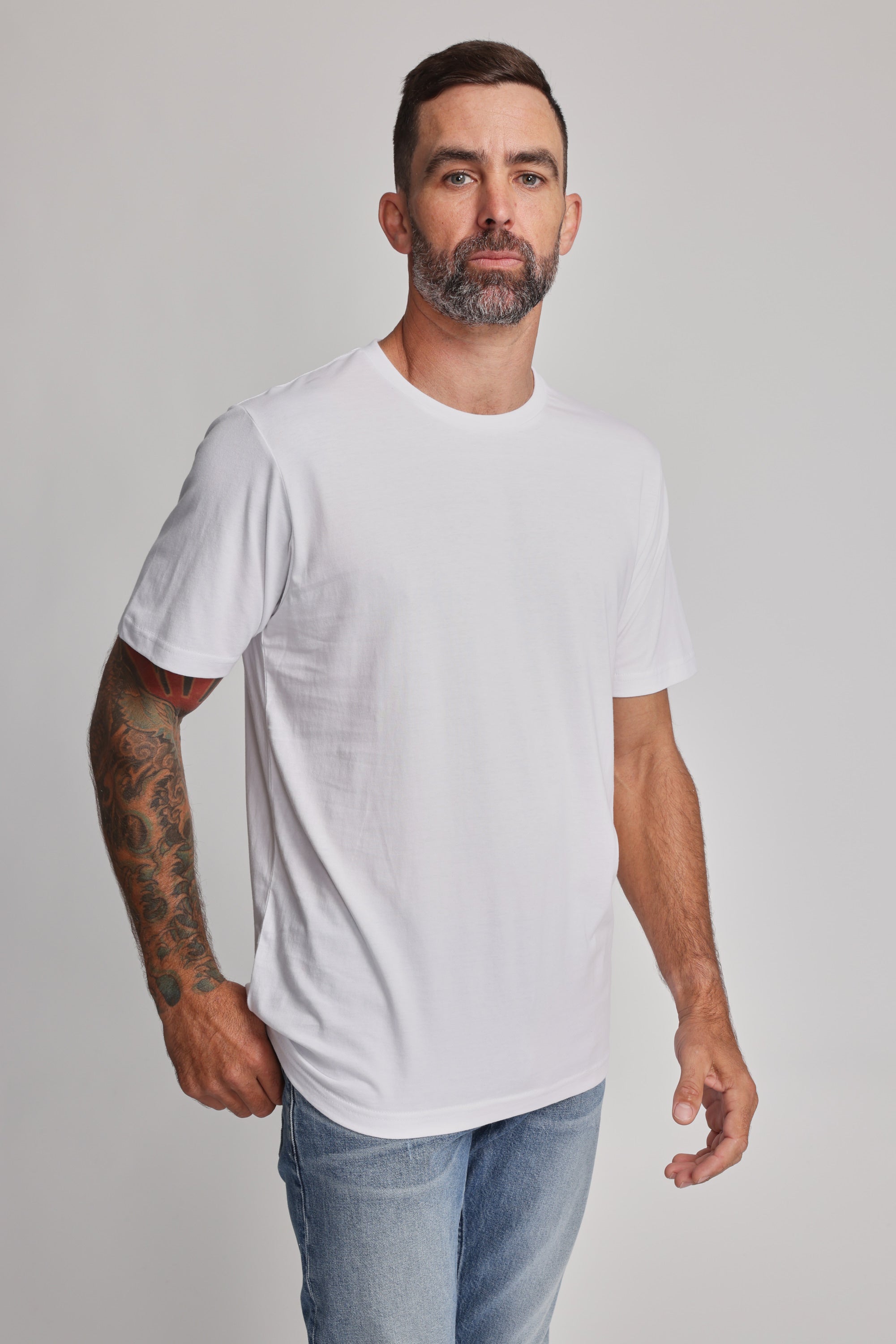 A man with short dark hair, a beard, and a tattooed right arm faces forward wearing the Barkers Organic Crew Tee in plain white with light blue jeans, standing against a light gray background.