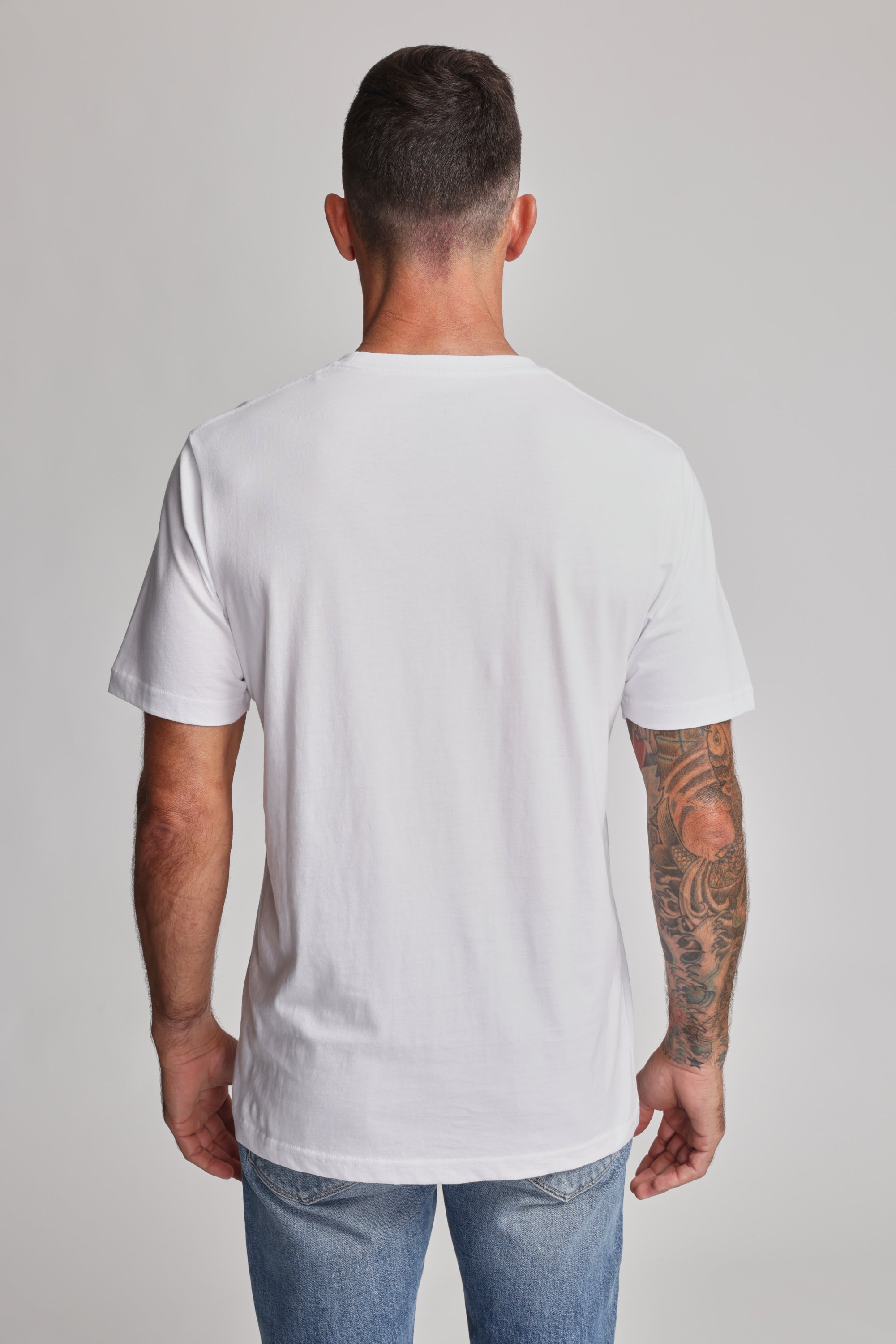 A man with short dark hair, wearing a plain white Barkers Organic Crew Tee and blue jeans, stands with his back to the camera. His right arm is tattooed, set against a neutral light gray background.