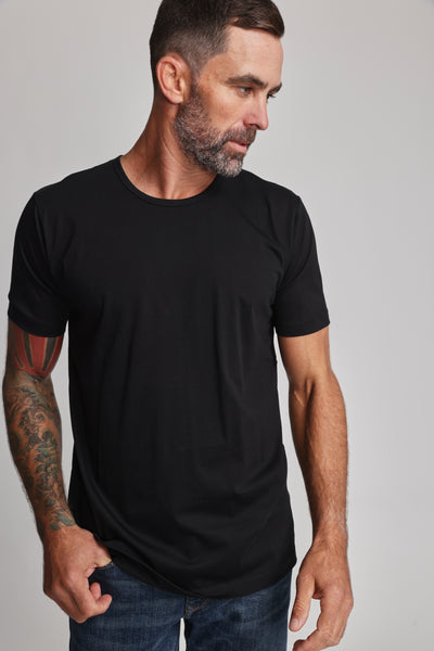 A man with short dark hair, a beard, and tattoos on his left arm wears the Barkers Linton Supima Tee in black with blue jeans. He looks down to his left against a plain light background.