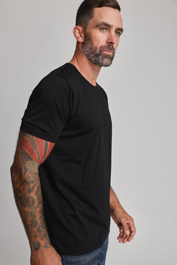 A man with short brown hair, a trimmed beard, and a tattoo sleeve on his left arm stands in Barkers' Linton Supima Tee and jeans against a plain light background, looking slightly to the side.