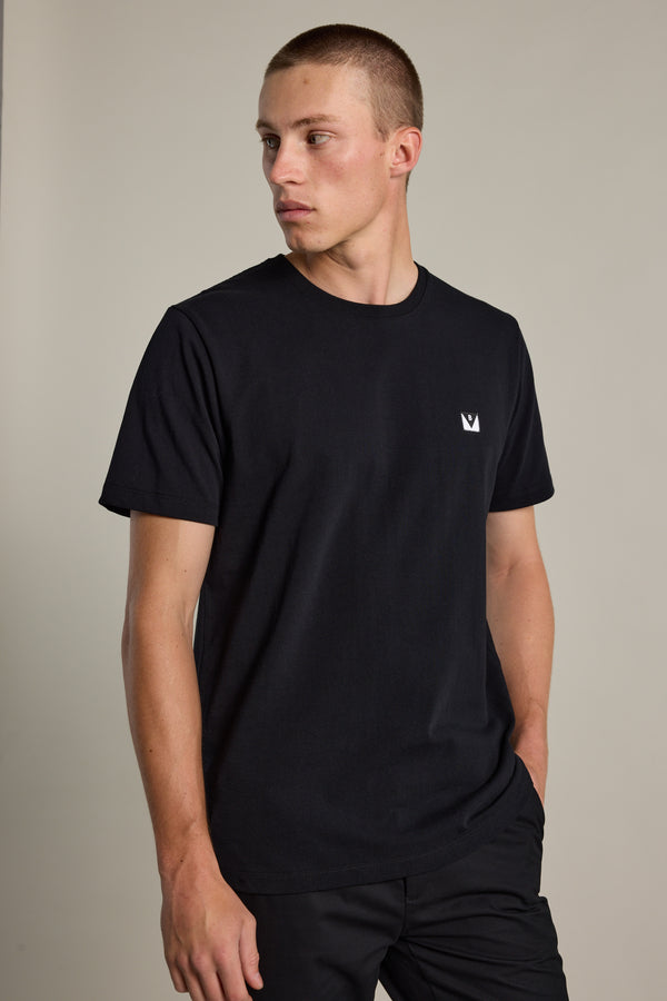 A young man with short hair wears the Barkers Newton Washed Tee, an oversized black box fit t-shirt featuring a small white crown logo on the left chest. He stands with one hand in his pocket, looking left against a plain light background.