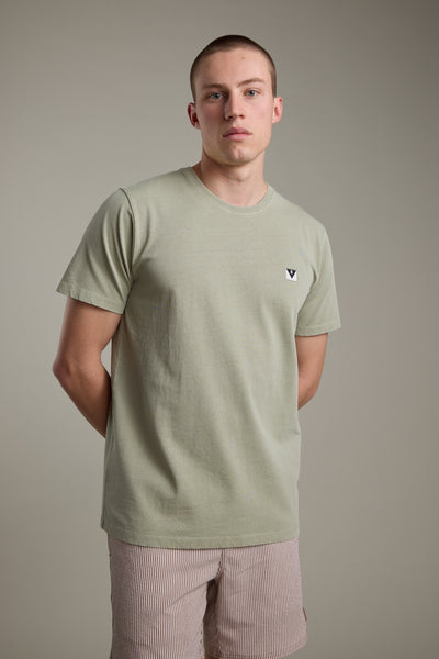 A young man with short hair stands against a plain background, wearing the Barkers Newton Washed Tee in light green featuring a small chest logo, paired with beige-striped shorts. His hands are behind his back as he looks directly at the camera.