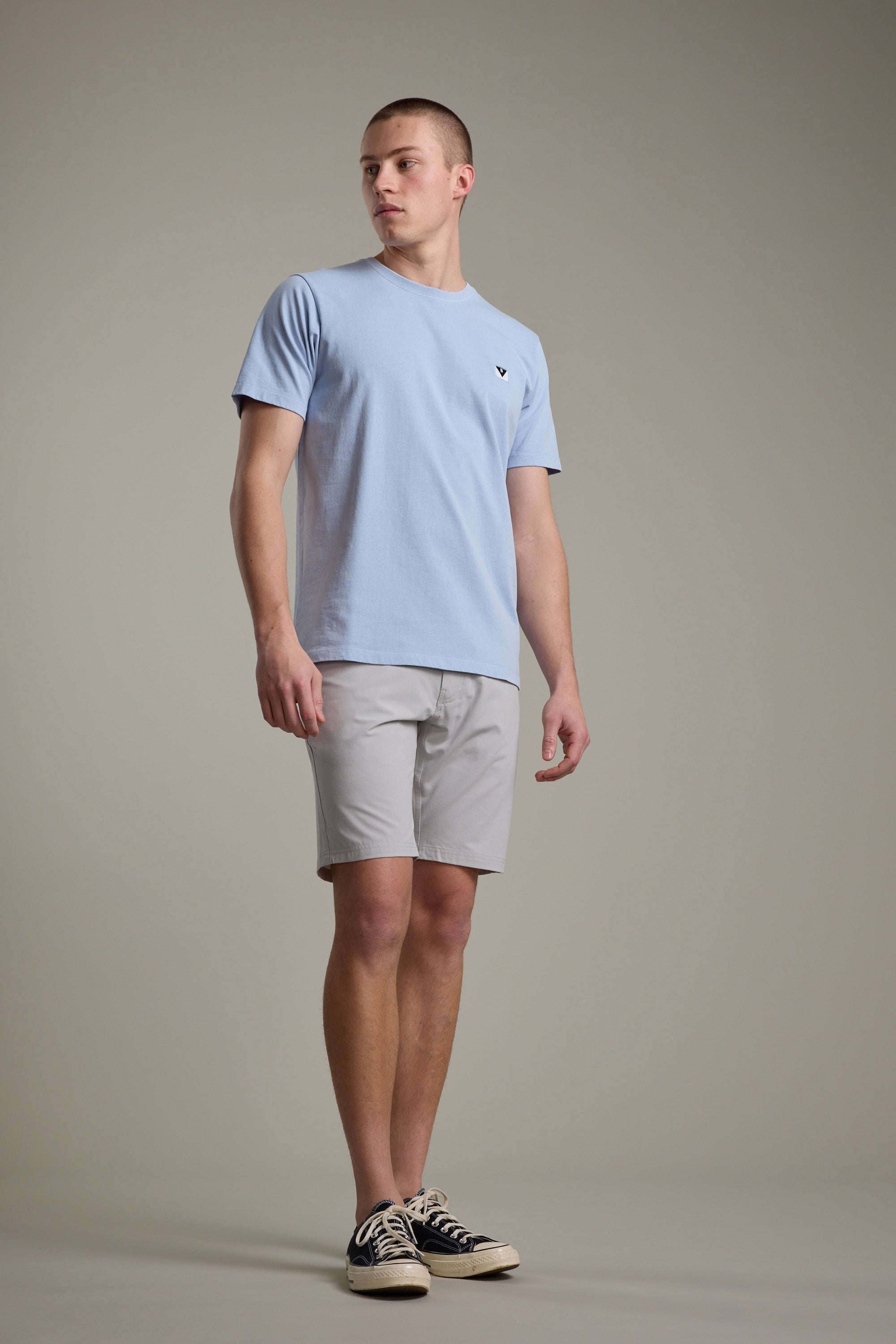 A person stands against a plain background wearing the Barkers Newton Washed Tee in light blue, paired with light gray shorts and black sneakers with white soles, looking slightly to the side with a neutral expression.