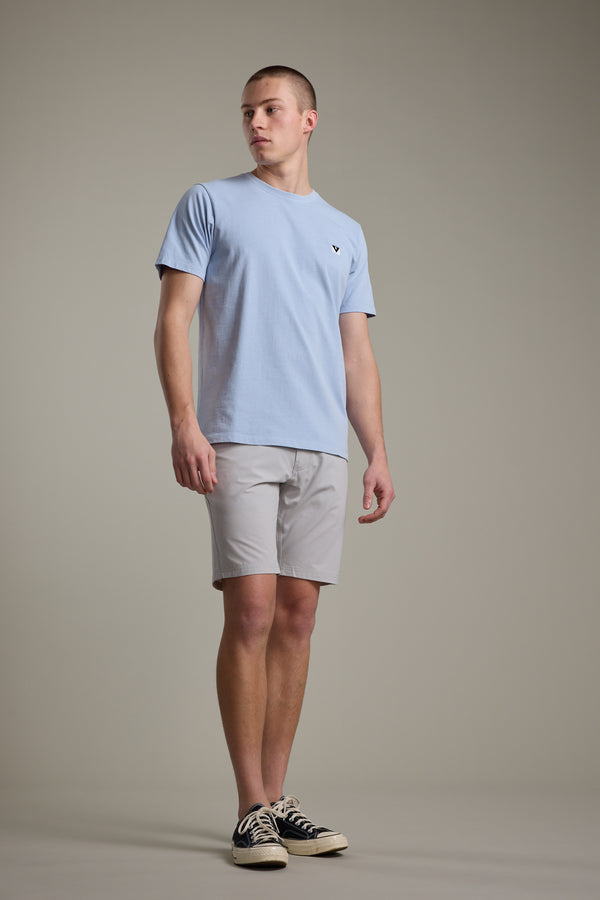 A person stands against a plain background wearing the Barkers Newton Washed Tee in light blue, paired with light gray shorts and black sneakers with white soles, looking slightly to the side with a neutral expression.
