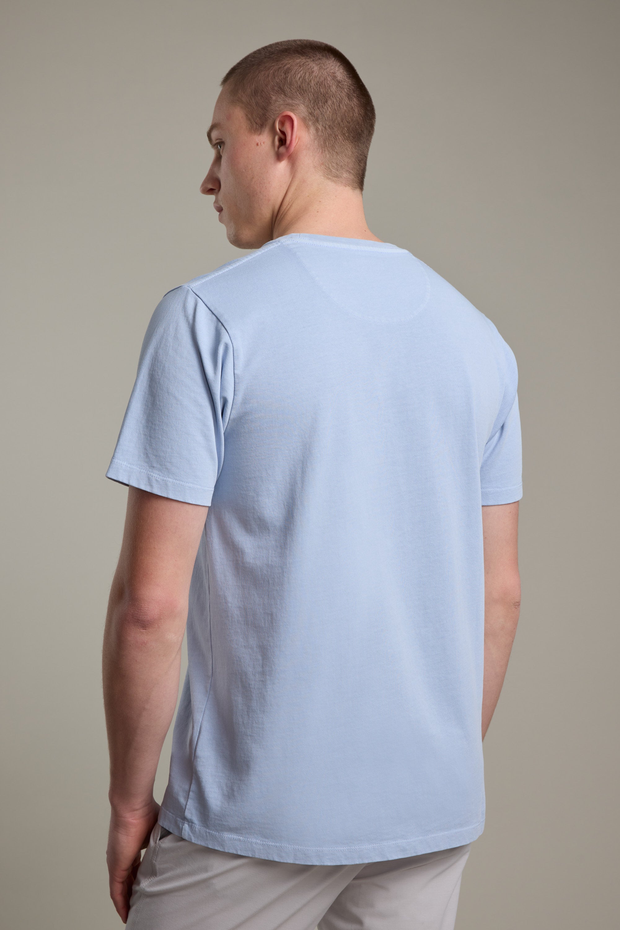 A man with short hair stands facing away from the camera, wearing the Barkers Newton Washed Tee and light-colored pants against a plain, neutral background.