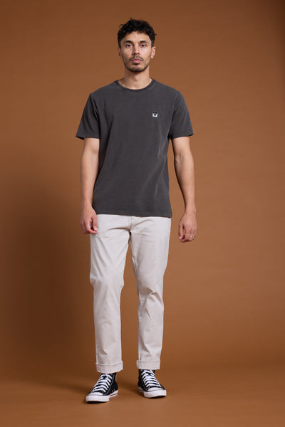 A man stands against a brown backdrop, wearing the Barkers Newton Washed Tee in charcoal gray, paired with light beige pants and black high-top sneakers. He faces forward with a neutral expression and relaxed posture.
