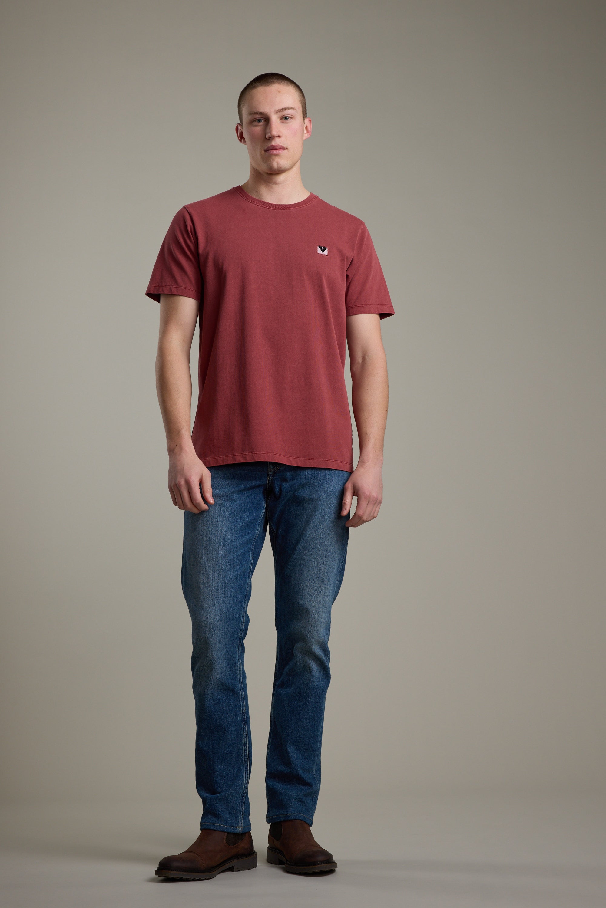 A young man with short hair and a neutral expression faces forward against a plain background, wearing the Barkers Newton Washed Tee in maroon, along with blue jeans and brown boots.