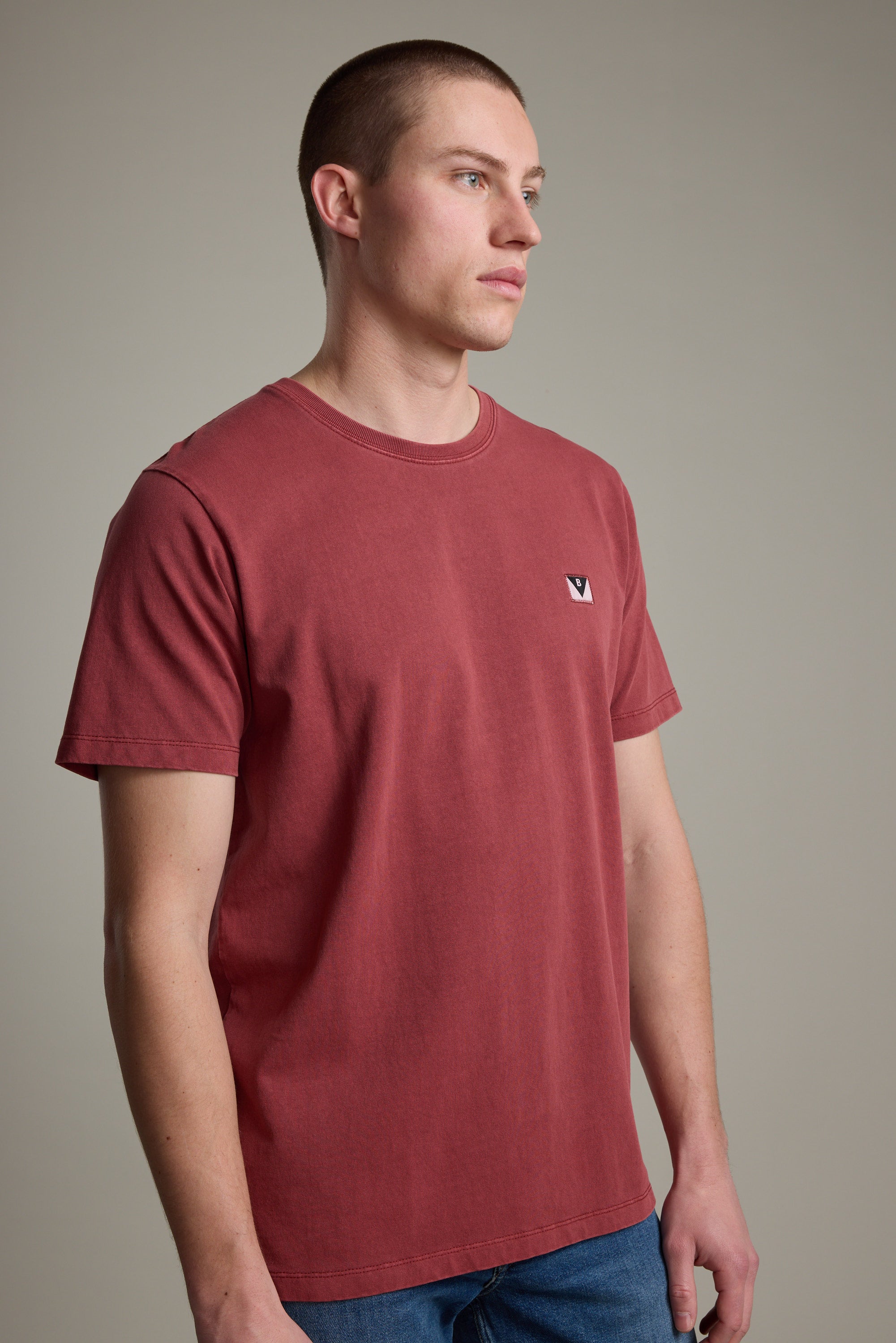 A man with short brown hair models the Barkers Newton Washed Tee in faded red, featuring a small white and black chest logo, as he stands against a plain gray background looking slightly to the side.