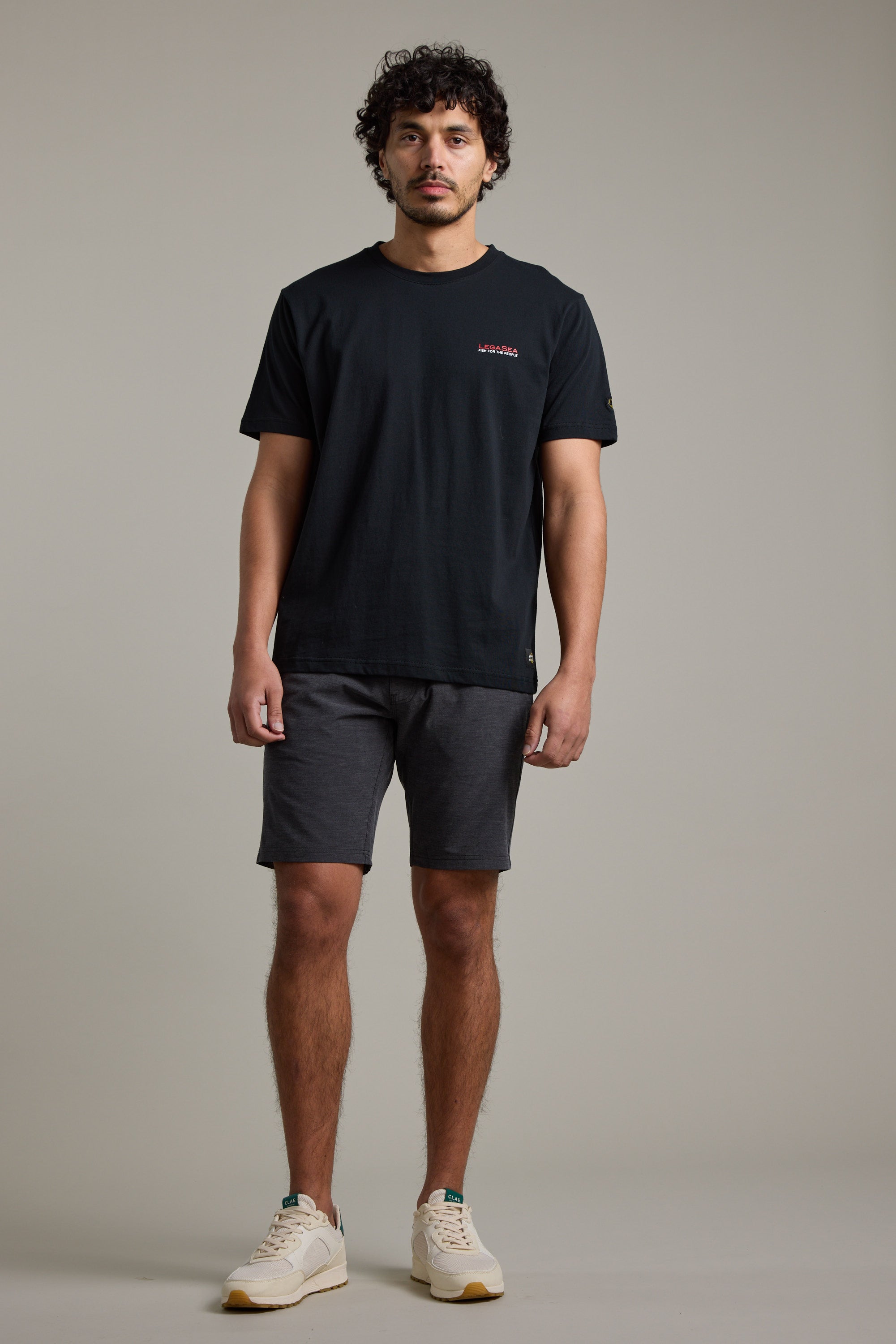 A man with curly hair stands against a plain background, wearing a Legasea Brand Tee by Legasea. Facing forward with a neutral expression and relaxed arms, he embodies support for New Zealand marine conservation.