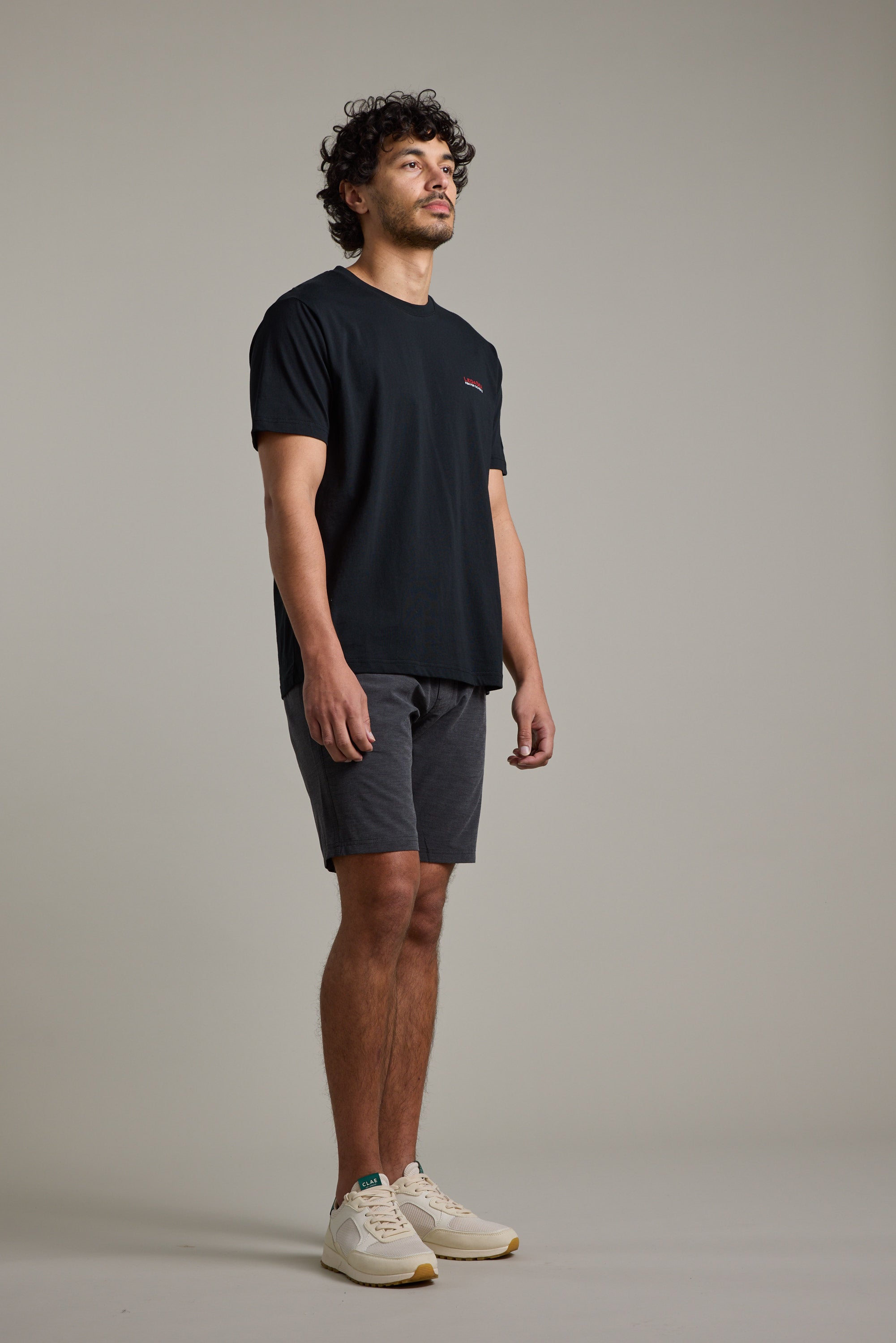 A man with curly hair stands against a plain background, wearing the Legasea Brand Tee—an organic cotton t-shirt by Legasea, supporting New Zealand marine conservation—paired with dark gray shorts and white sneakers. He faces slightly right, expression neutral.