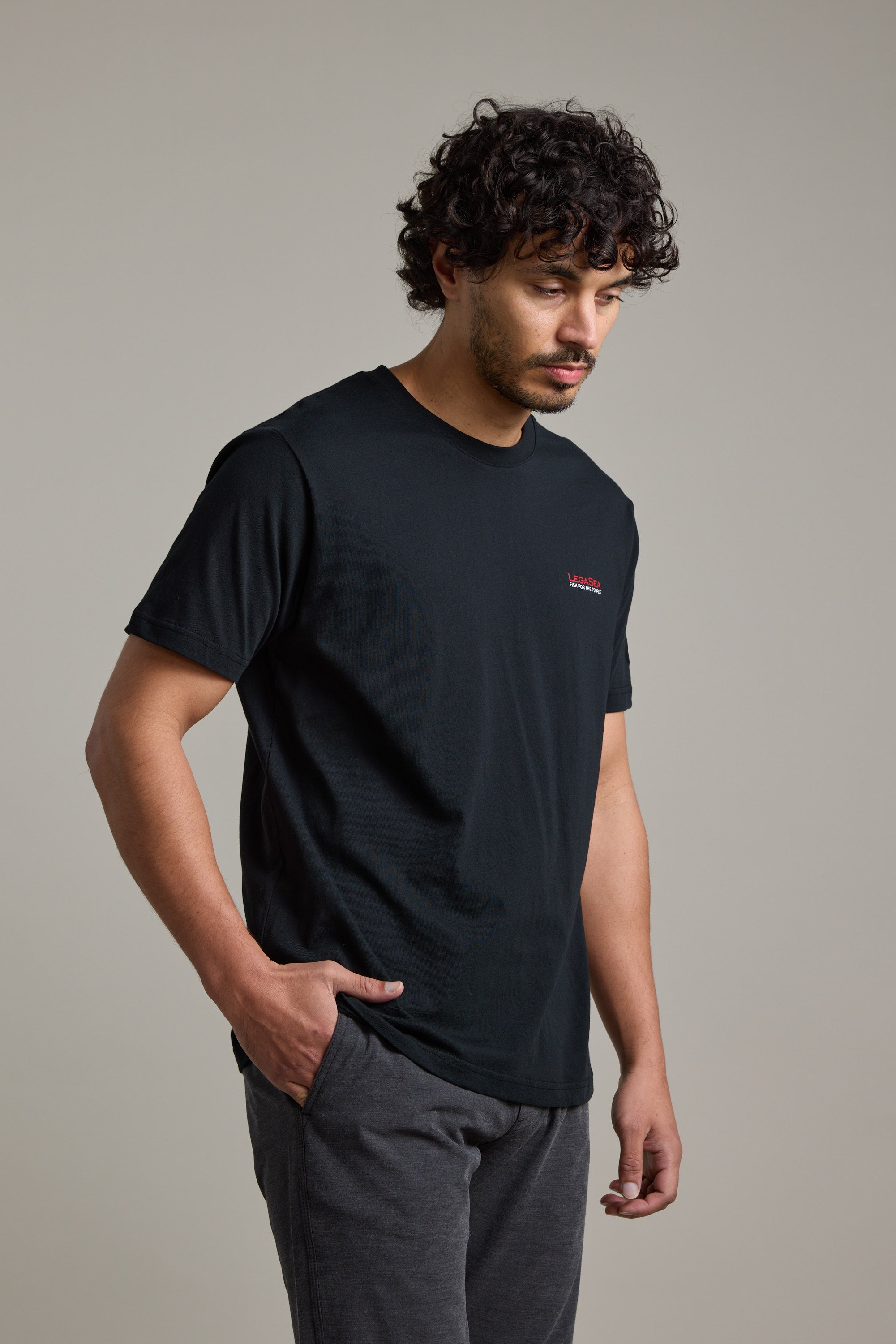A man with curly hair wears a Legasea Brand Tee by Legasea—an organic cotton t-shirt supporting marine conservation in New Zealand. He stands against a plain background, looking down and to the side with one hand in his pocket.