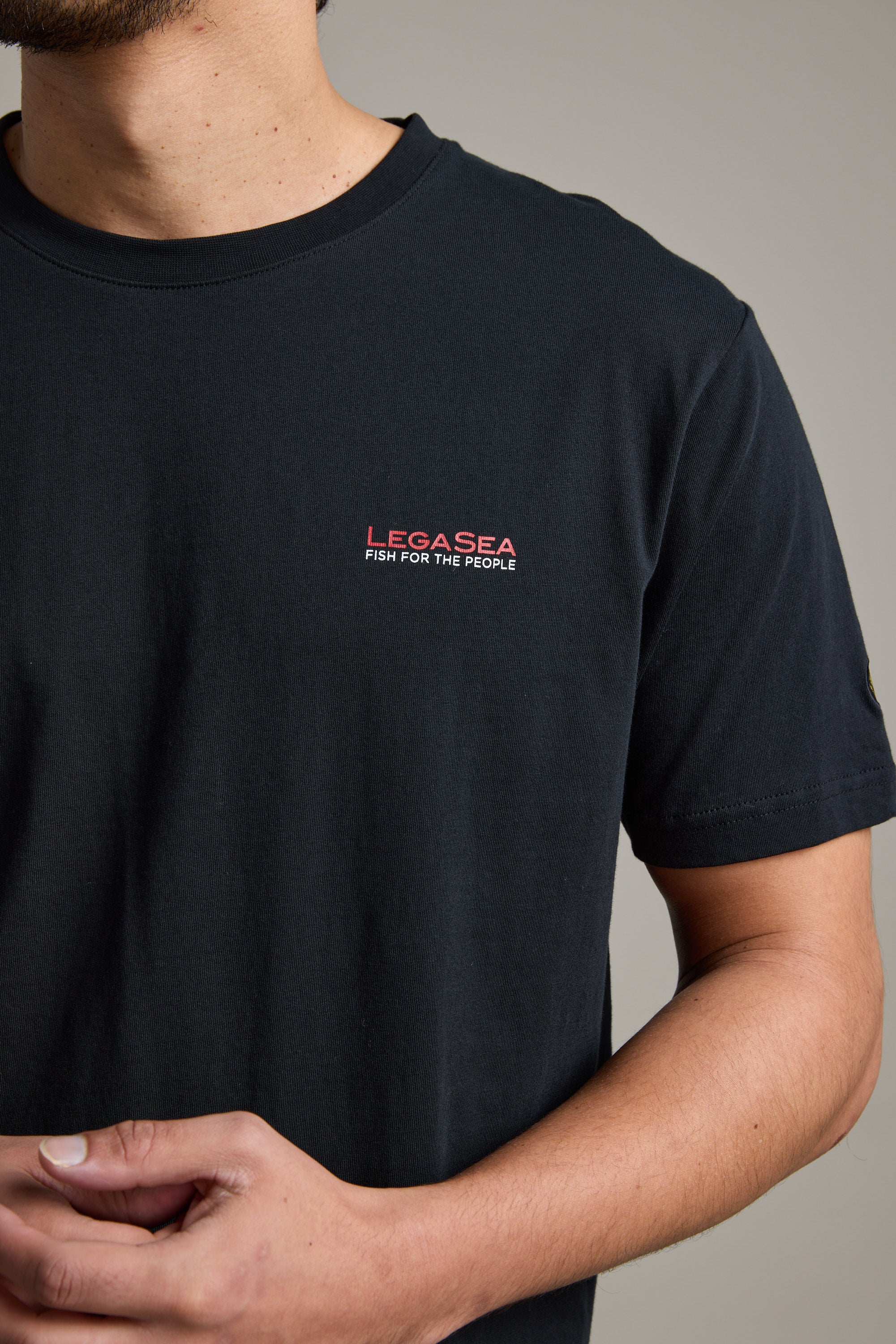 A person wears a Legasea Brand Tee, an organic cotton t-shirt by Legasea with “LECASEA FISH FOR THE PEOPLE” in red and white on the left chest, supporting NZ marine conservation. Face not shown; hands are lightly clasped.