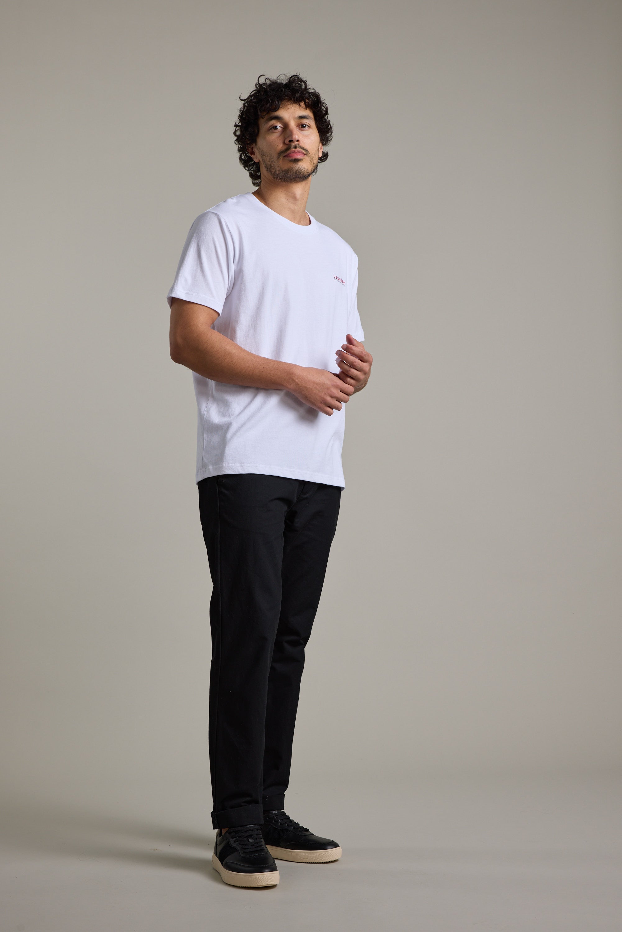 A man with curly hair stands against a plain background, wearing a Legasea Brand Tee in white, paired with black pants and black sneakers with white soles. He gazes slightly upward with a neutral expression.