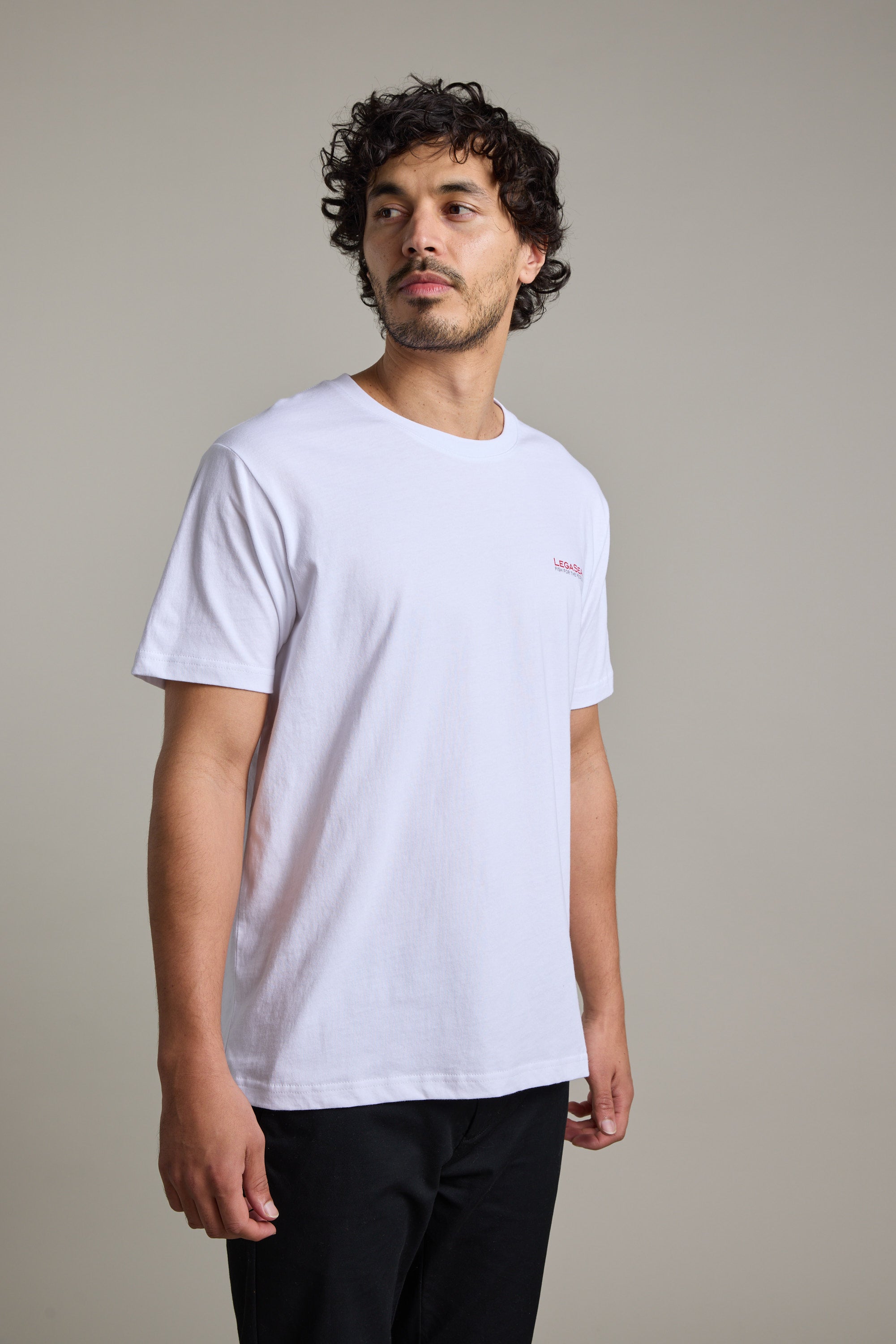 A man with curly hair and facial hair wears the Legasea Brand Tee by Legasea in white with black pants, standing against a neutral background and looking slightly to his left.