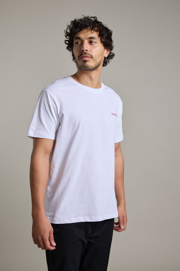 A man with curly hair and facial hair wears the Legasea Brand Tee by Legasea in white with black pants, standing against a neutral background and looking slightly to his left.