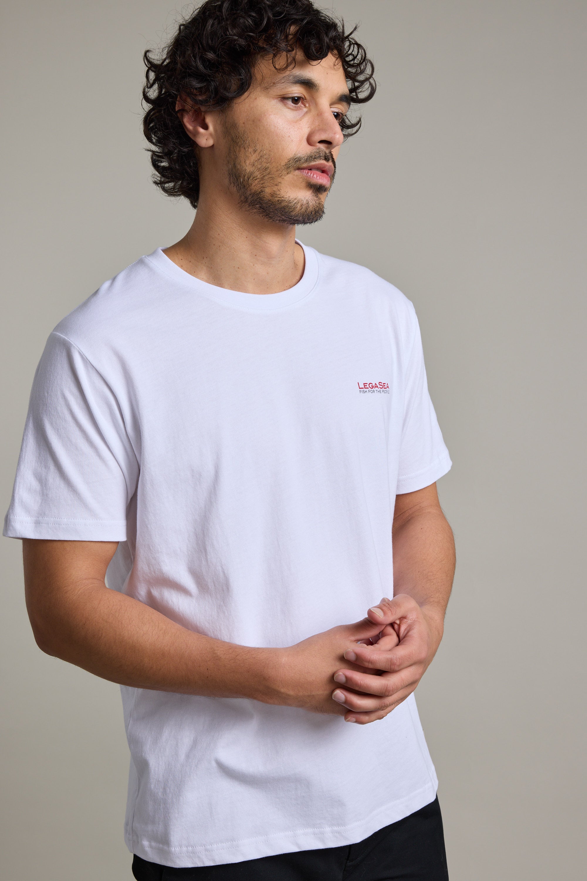 A man with curly hair and light facial hair wears the Legasea Brand Tee by Legasea and black pants, standing against a neutral background with his hands lightly clasped in front of him.