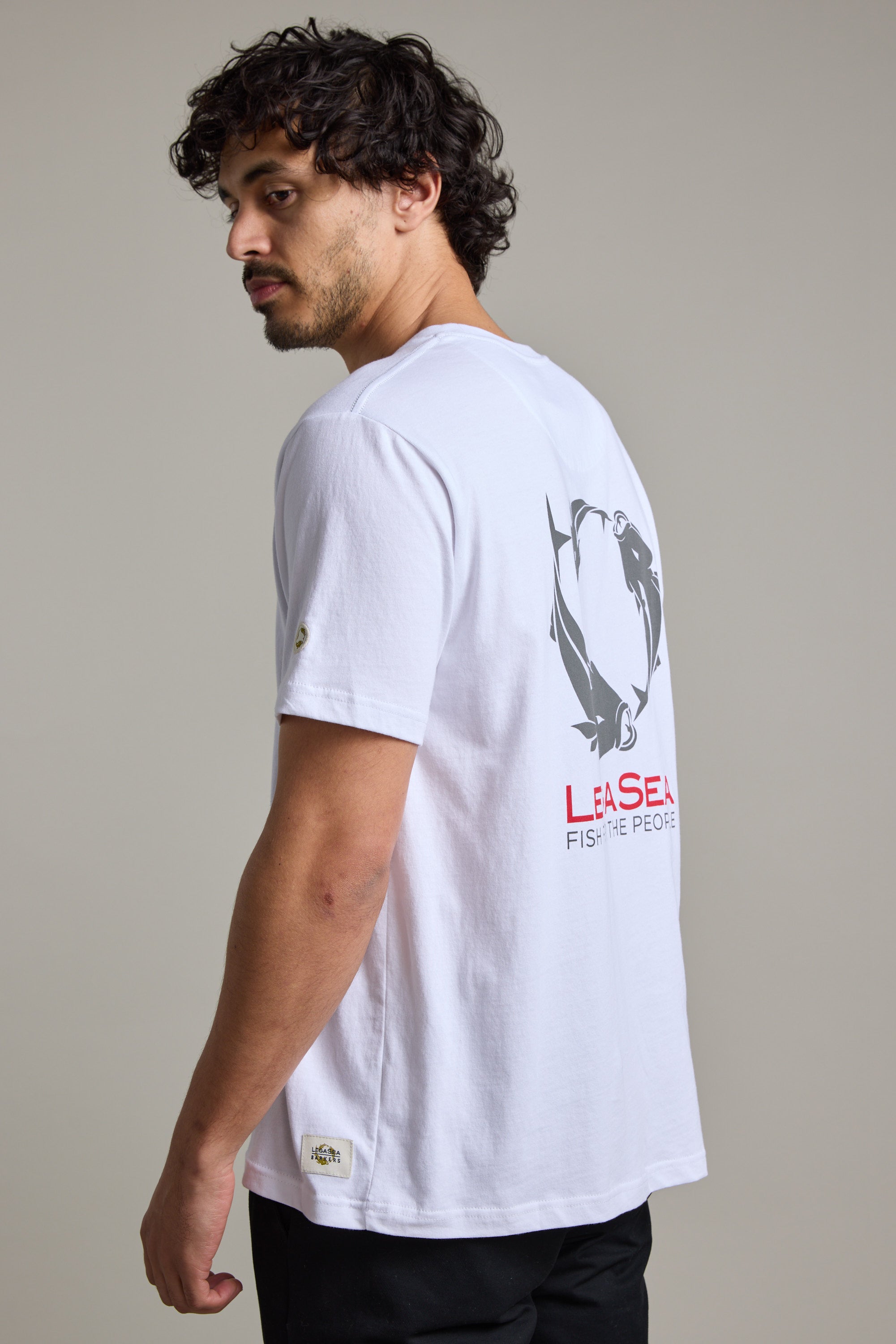 A man with curly hair and a beard wears a Legasea Brand Tee made of organic cotton, featuring a black fish graphic and bold red "LEA SEAL FISH THE PEOPLE" text on the back. He looks over his shoulder against a plain background.