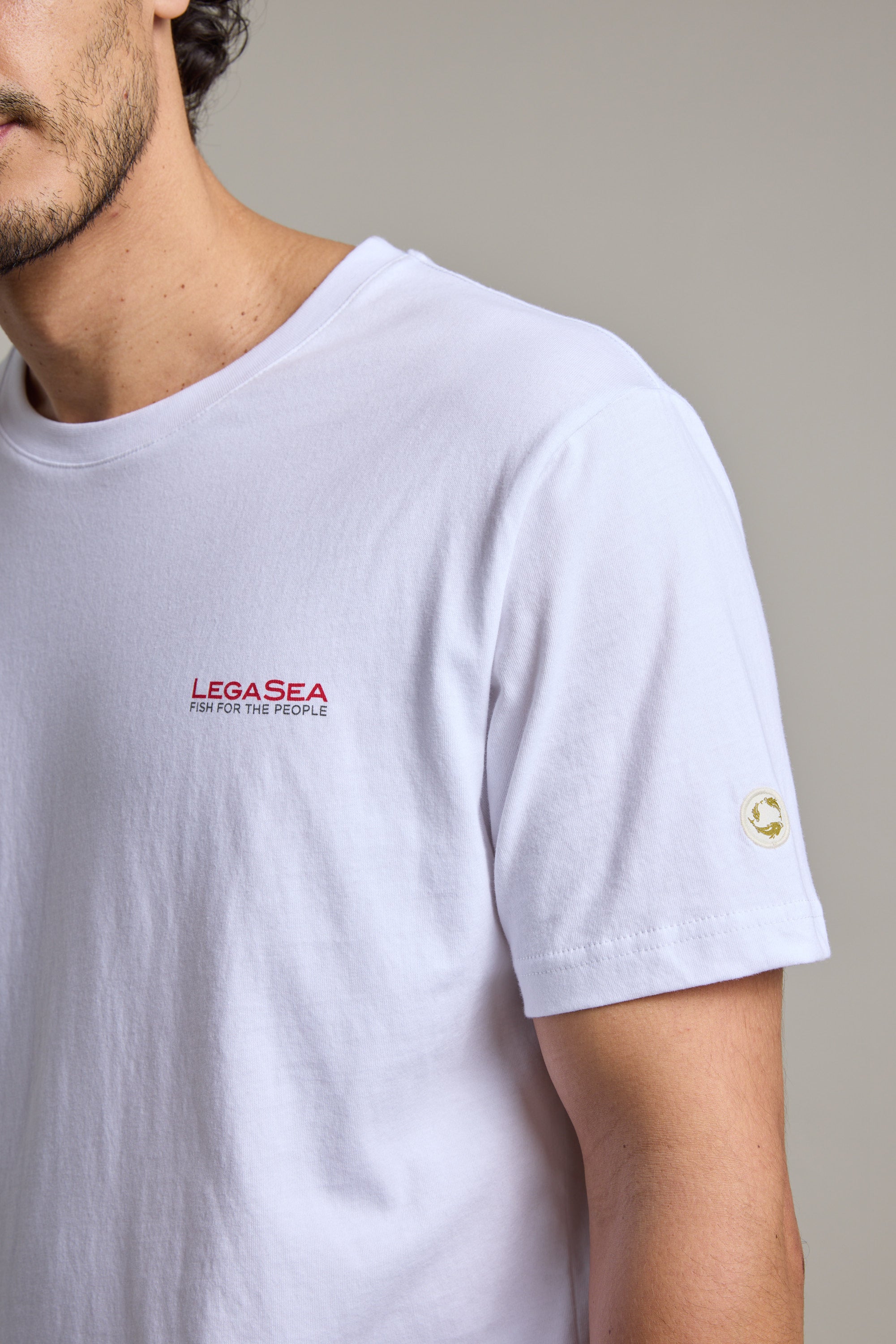 A person wears the Legasea Brand Tee—made from organic cotton, featuring bold red “LEGASEA FISH FOR THE PEOPLE” text on the chest and a small circular Legasea logo on the sleeve. Only their upper torso and part of the face are visible.