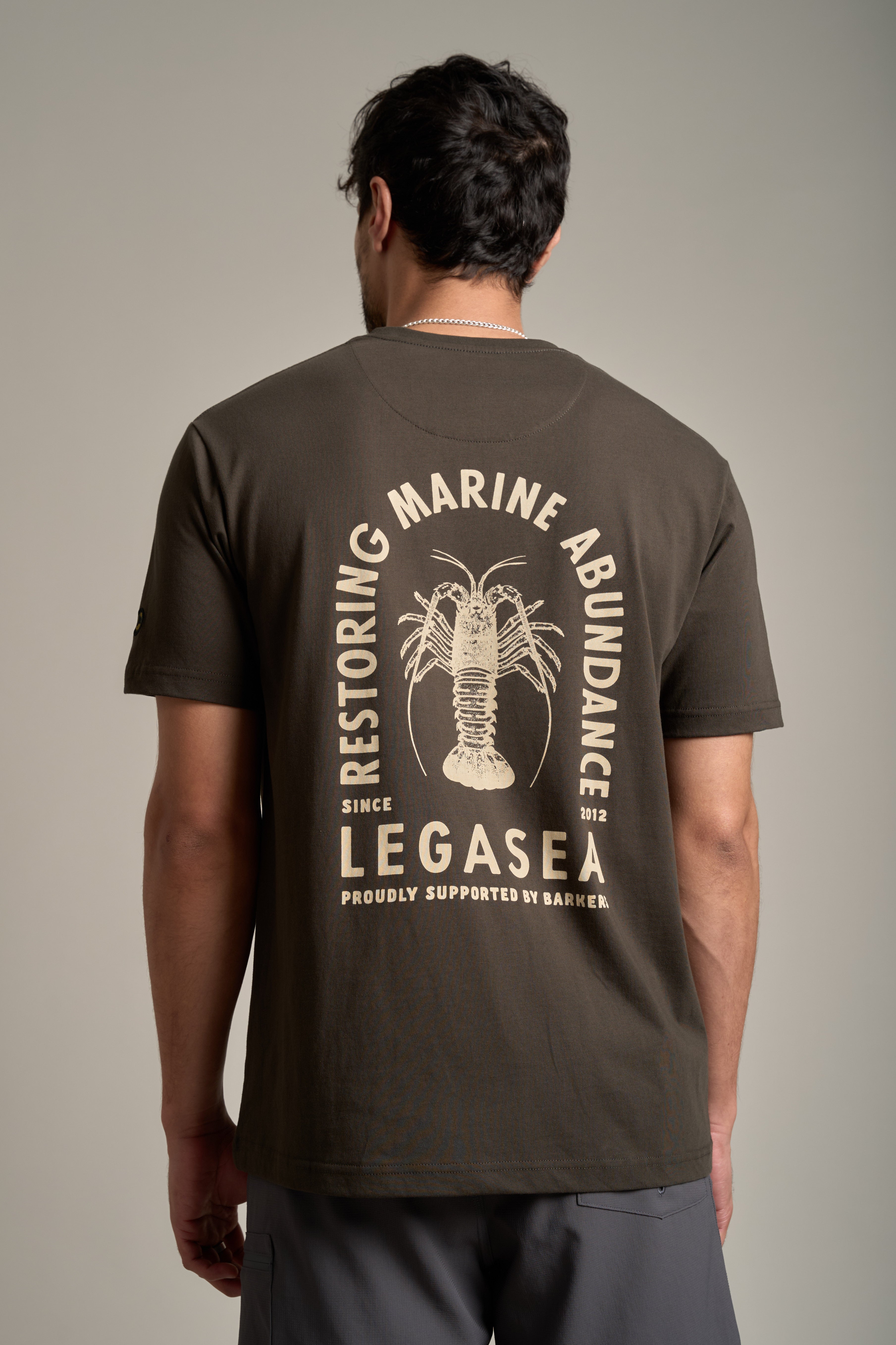 A man with short dark hair faces away, wearing the Legasea Crayfish Print Tee—an organic cotton t-shirt featuring a crayfish graphic and “Restoring Marine Abundance, Legasea. Proudly Supported by Barkers.”.