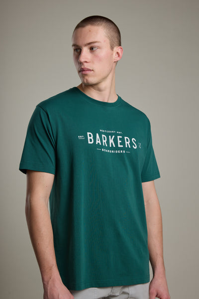 A person in the Barkers Boardriders Logo Tee, a green organic cotton shirt with logo and text on the front, stands against a plain light gray background looking to the side.
