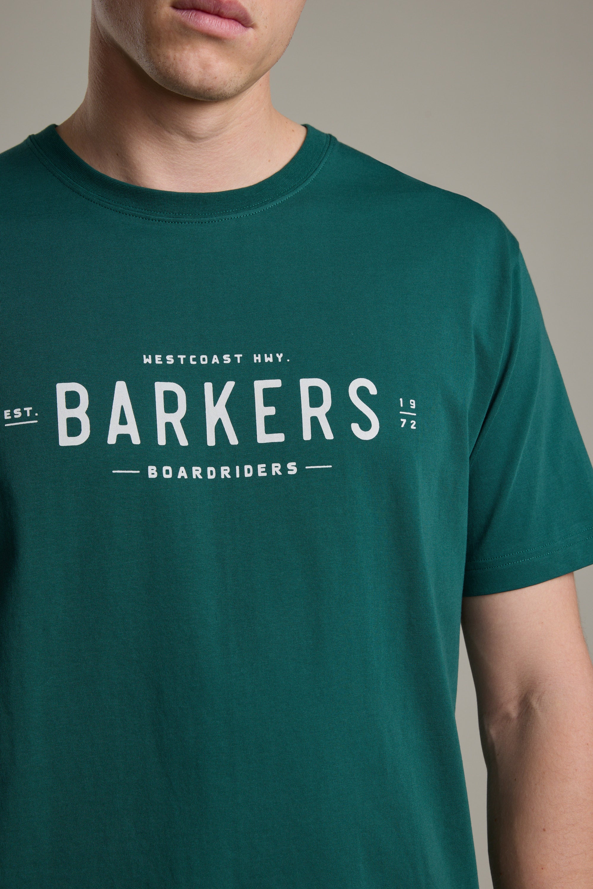 A person wears the Barkers Boardriders Logo Tee in dark green organic cotton, featuring the Westcoast Hwy. logo and "Est. 1972" on the front. The photo highlights this everyday essential by focusing on the upper torso and part of their face.