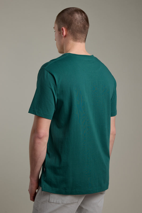 A person with short hair, facing away from the camera, wears a green Barkers Boardriders Logo Tee and light gray pants against a plain, neutral background.