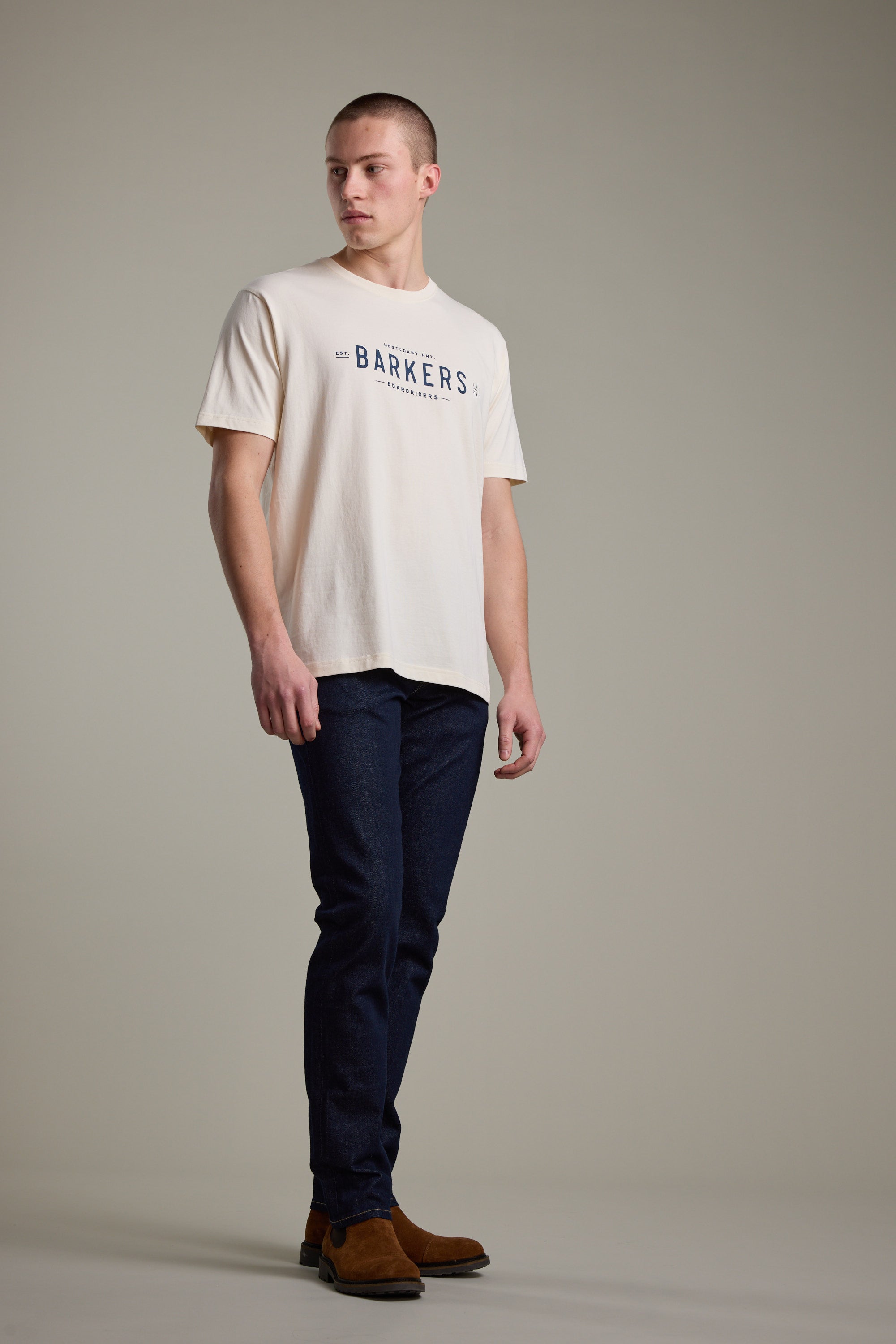 A man stands in a studio wearing a Barkers Boardriders Logo Tee, dark blue jeans, and brown suede boots, looking off to the side with a neutral expression against a plain background.