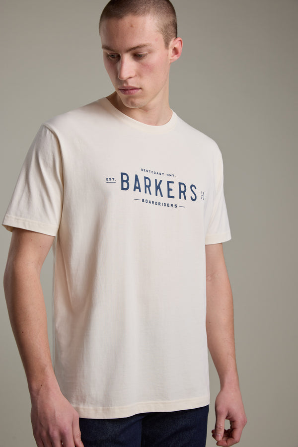 A person wears the Barkers Boardriders Logo Tee in light beige organic cotton, featuring a blue Barkers Boardriders logo on the front, standing against a plain background and looking down to the side.