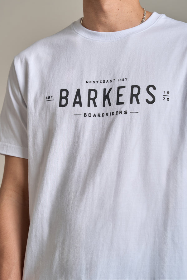 A person wears the Barkers Boardriders Logo Tee, an organic cotton essential featuring the Barkers logo and "WESTCOAST HWY. BARKERS BOARD RIDERS EST. 1972" printed in black across the chest; only their upper body shows.