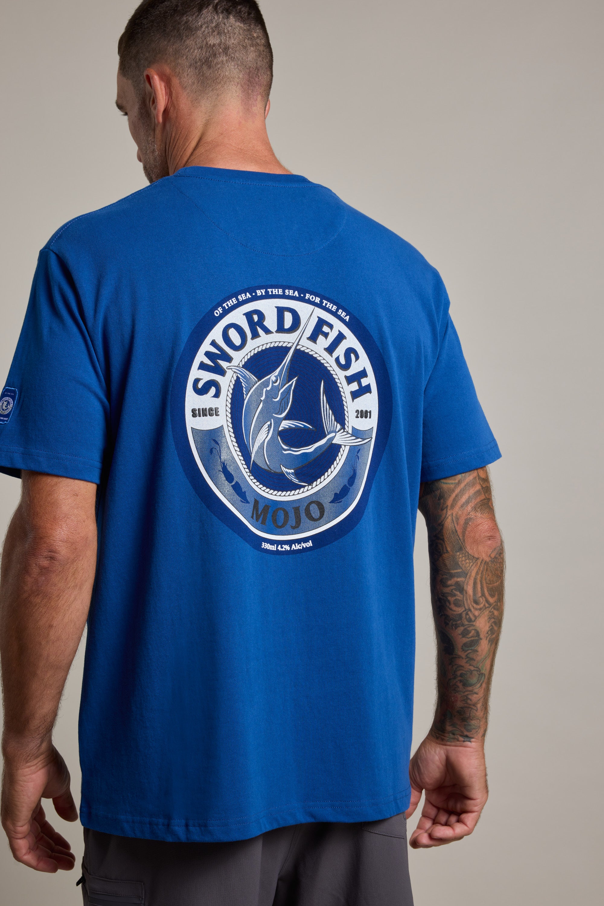 A man wears a blue Swordfish Mojo Logo Tee by Swordfish Mojo, featuring a large circular logo and fish graphic on the back. His tattooed right arm stands out against the plain background.