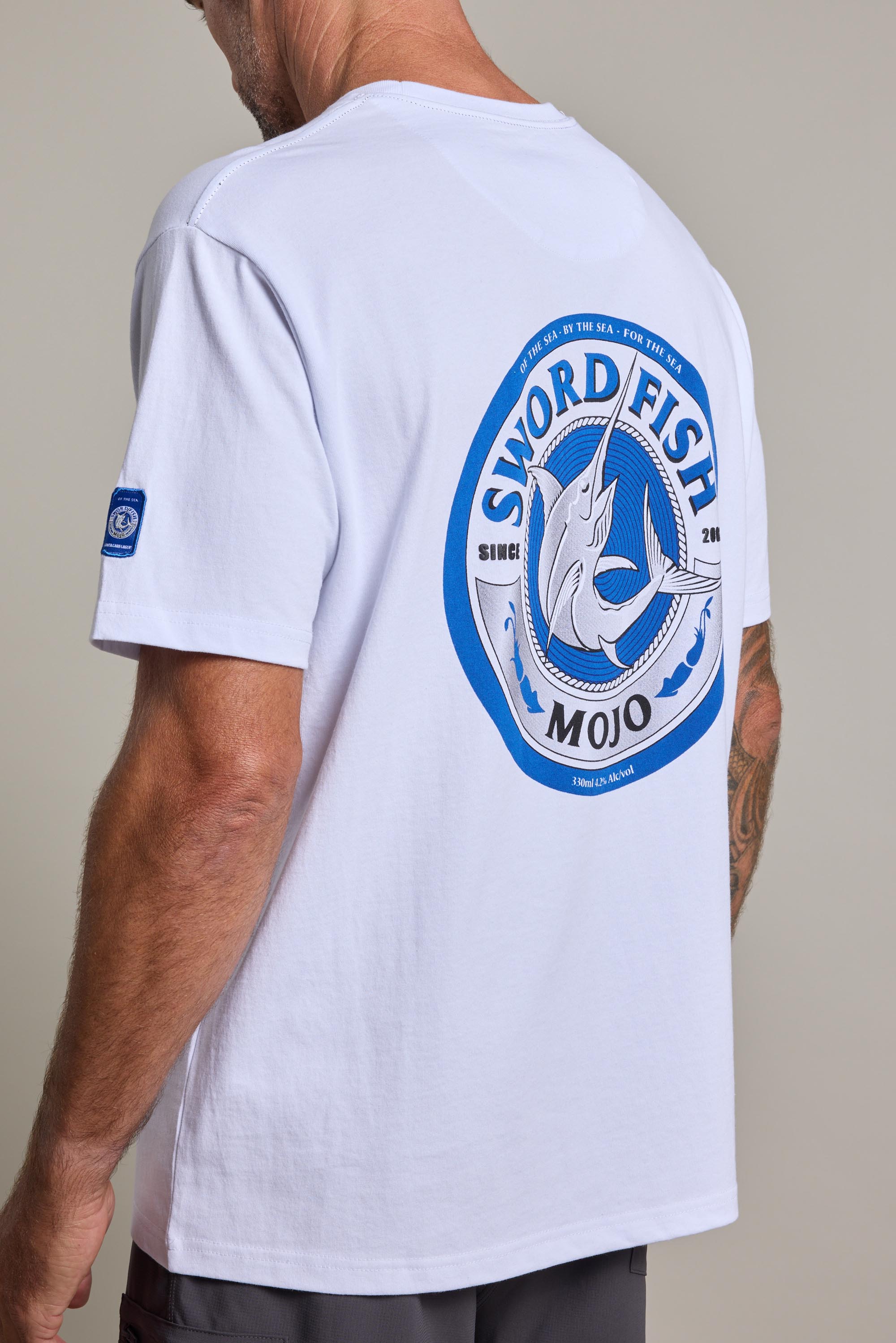 A man shown from behind wears the Swordfish Mojo Logo Tee by Swordfish Mojo, featuring a large blue swordfish graphic and circular text on the back—ideal merch for fans. A tattoo is visible on his arm.