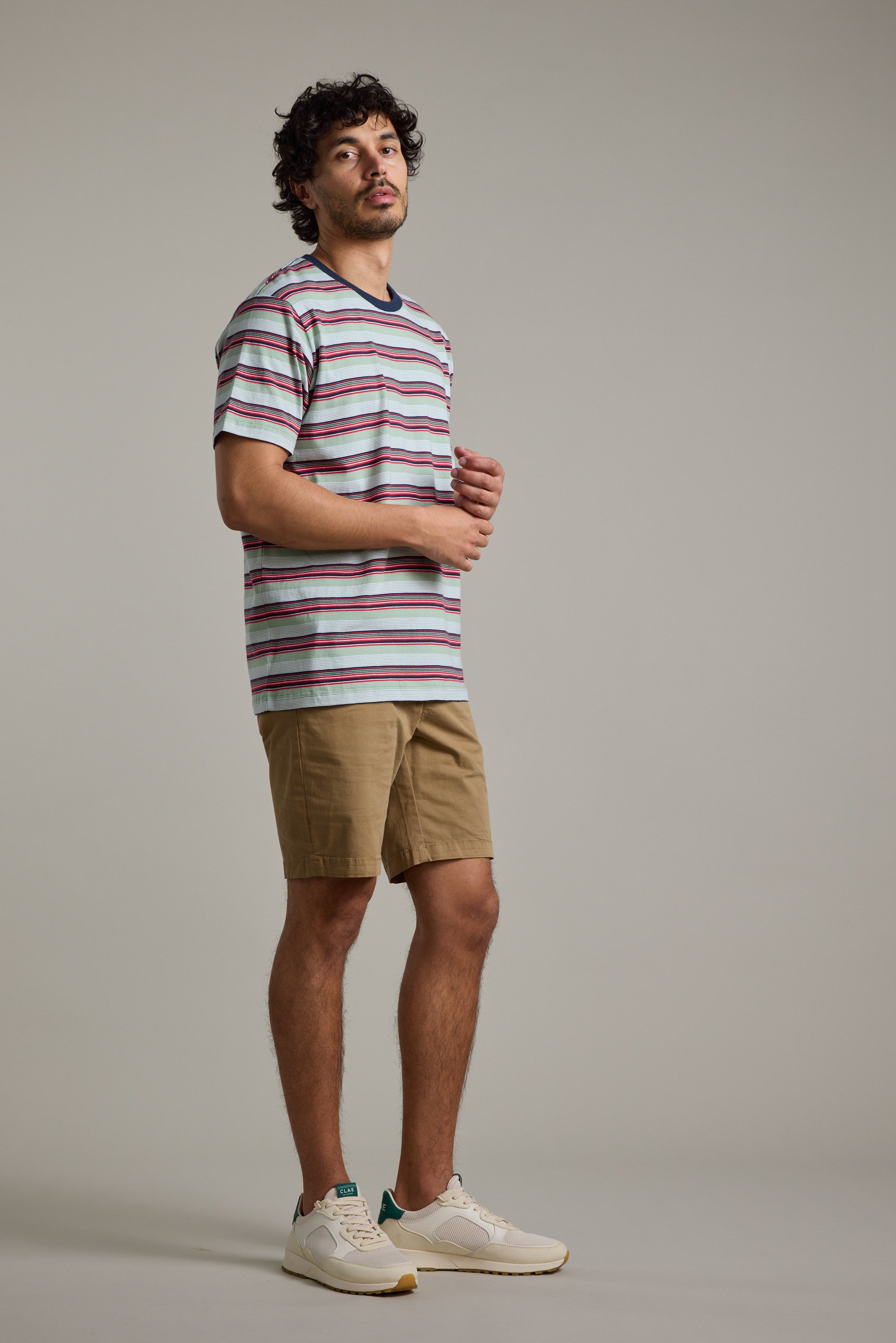 A man with curly hair stands in a studio wearing the Barkers Alvin Stripe Tee, khaki shorts, and white sneakers. He looks slightly to the side with a neutral expression against a plain background.