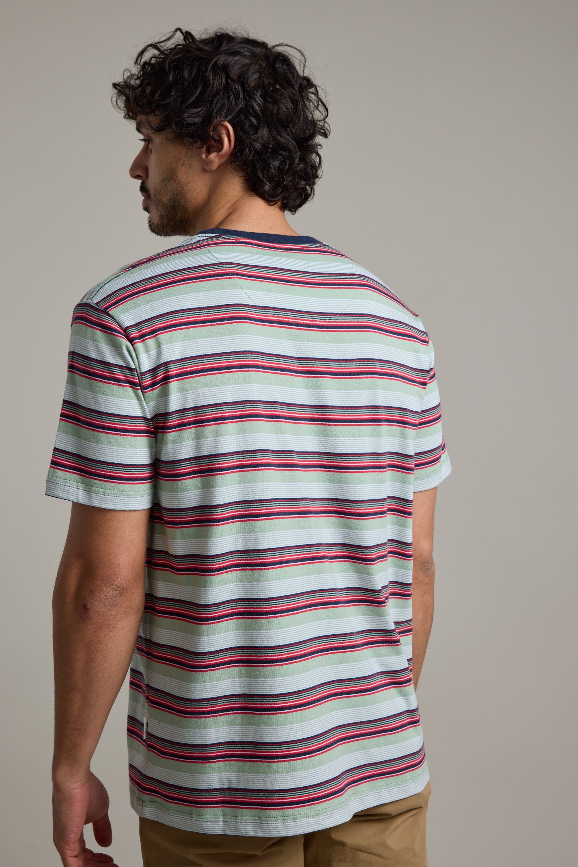 A man with curly dark hair, seen from behind, wears the Barkers Alvin Stripe Tee—an organic cotton crew neck shirt with red, blue, and black stripes—paired with tan pants against a plain neutral background.
