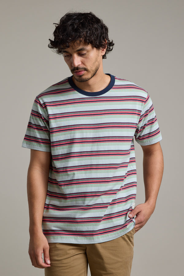 A man with wavy dark hair and facial hair wears the Barkers Alvin Stripe Tee—an organic cotton, light gray crewneck with horizontal red, blue, and black stripes—paired with tan pants against a plain gray background.