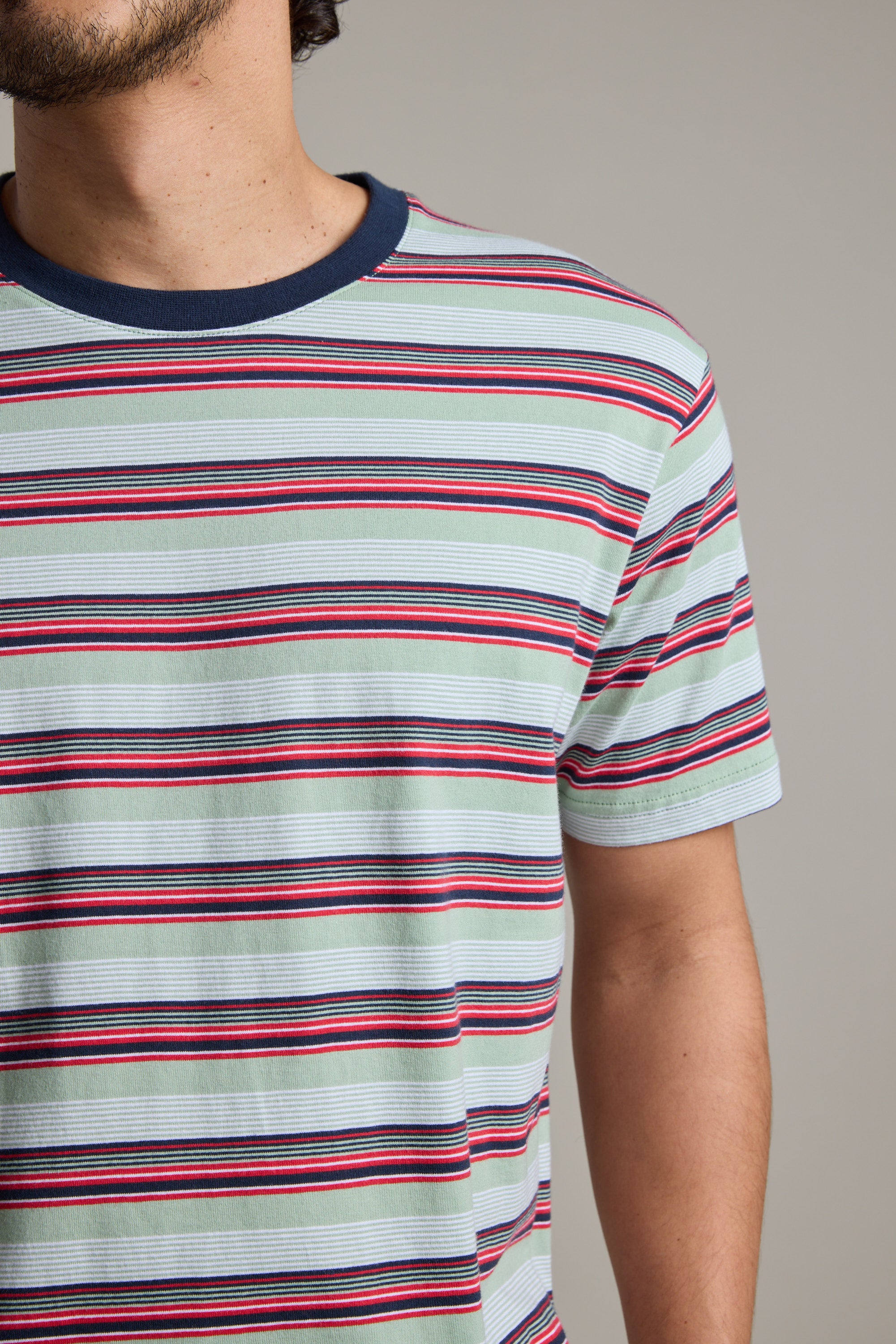 A person wearing the Barkers Alvin Stripe Tee—an organic cotton, short-sleeved shirt with red, black, and white horizontal stripes and a dark blue crew neck—shown from shoulders to waist, facing slightly left.