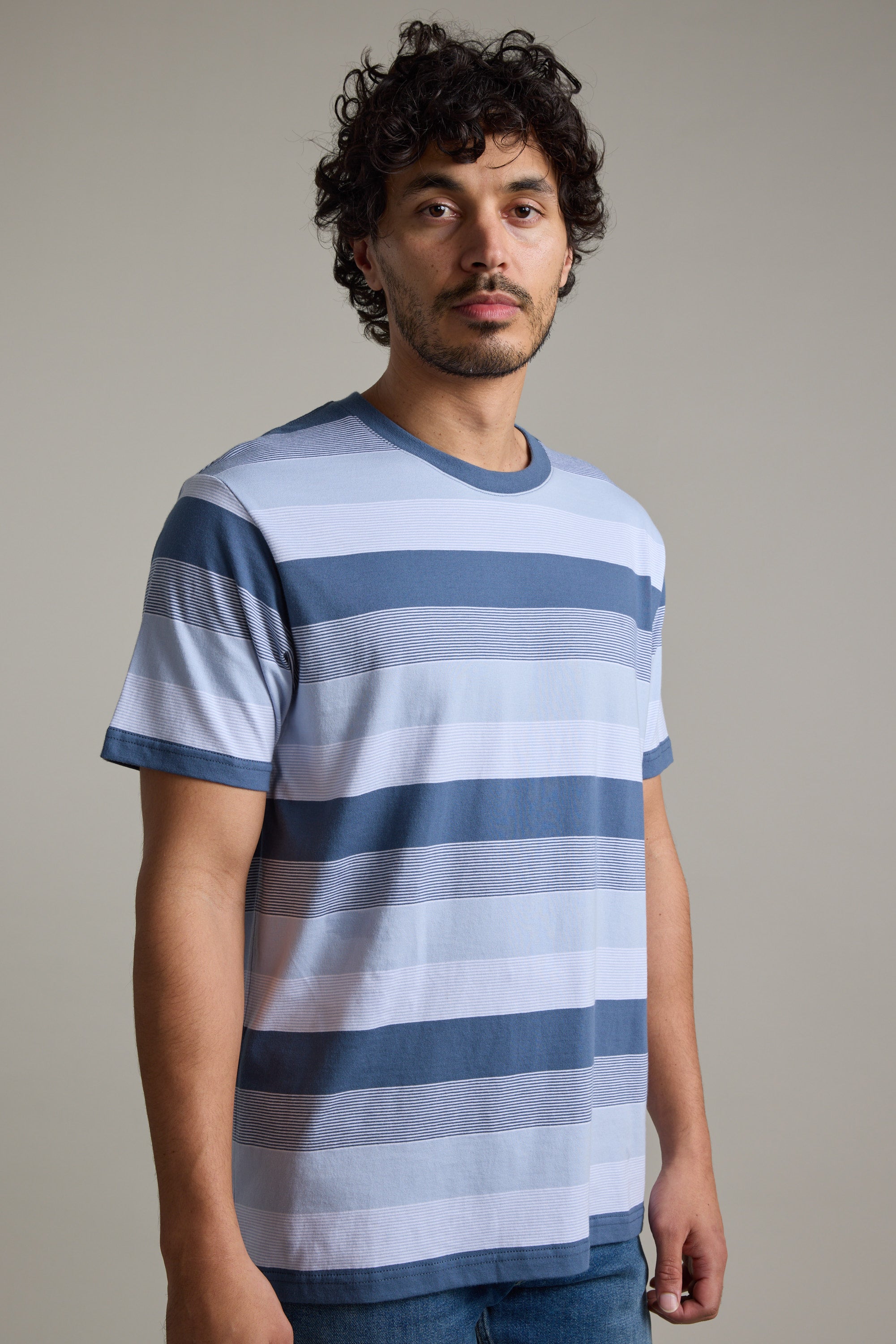 A man with curly dark hair and a short beard is wearing the Barkers Fitzroy Stripe Tee with blue jeans, standing in front of a plain, light-colored background.