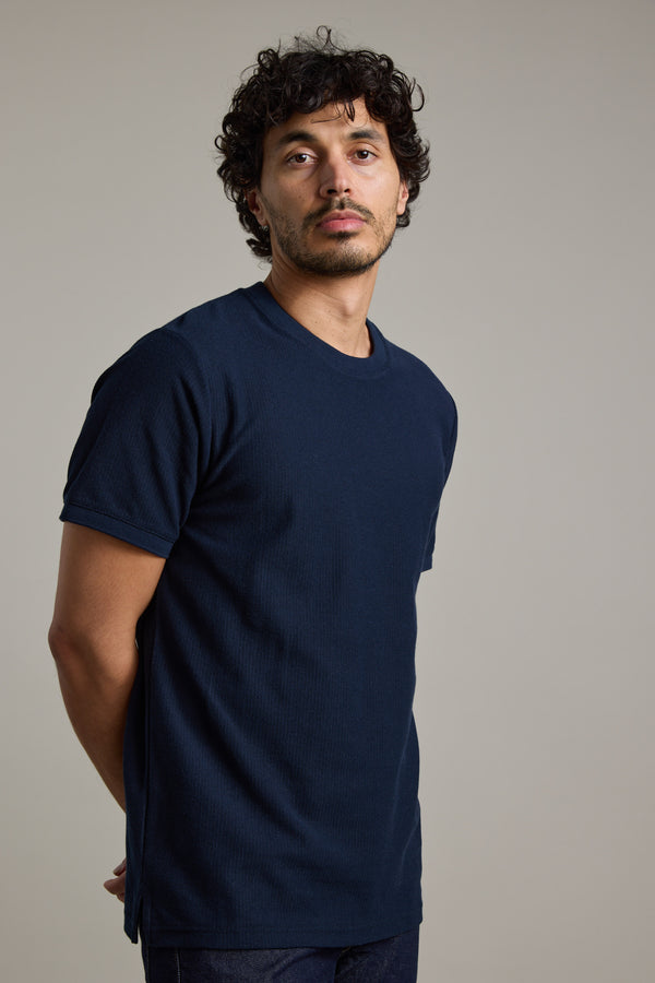 A man with curly dark hair and light facial hair stands against a gray background, wearing the Barkers Signature Knit Tee in navy blue. His arms are behind his back as he looks directly at the camera.