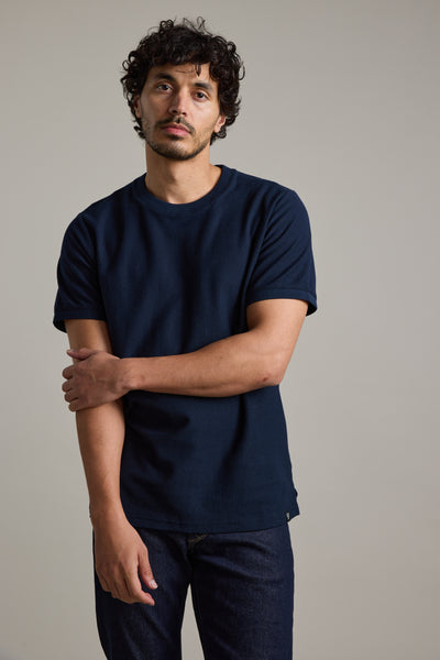 A man with curly dark hair and slight facial hair stands against a plain background, wearing the Barkers Signature Knit Tee in dark blue and dark jeans, with one arm crossed over to hold his other arm above the elbow.