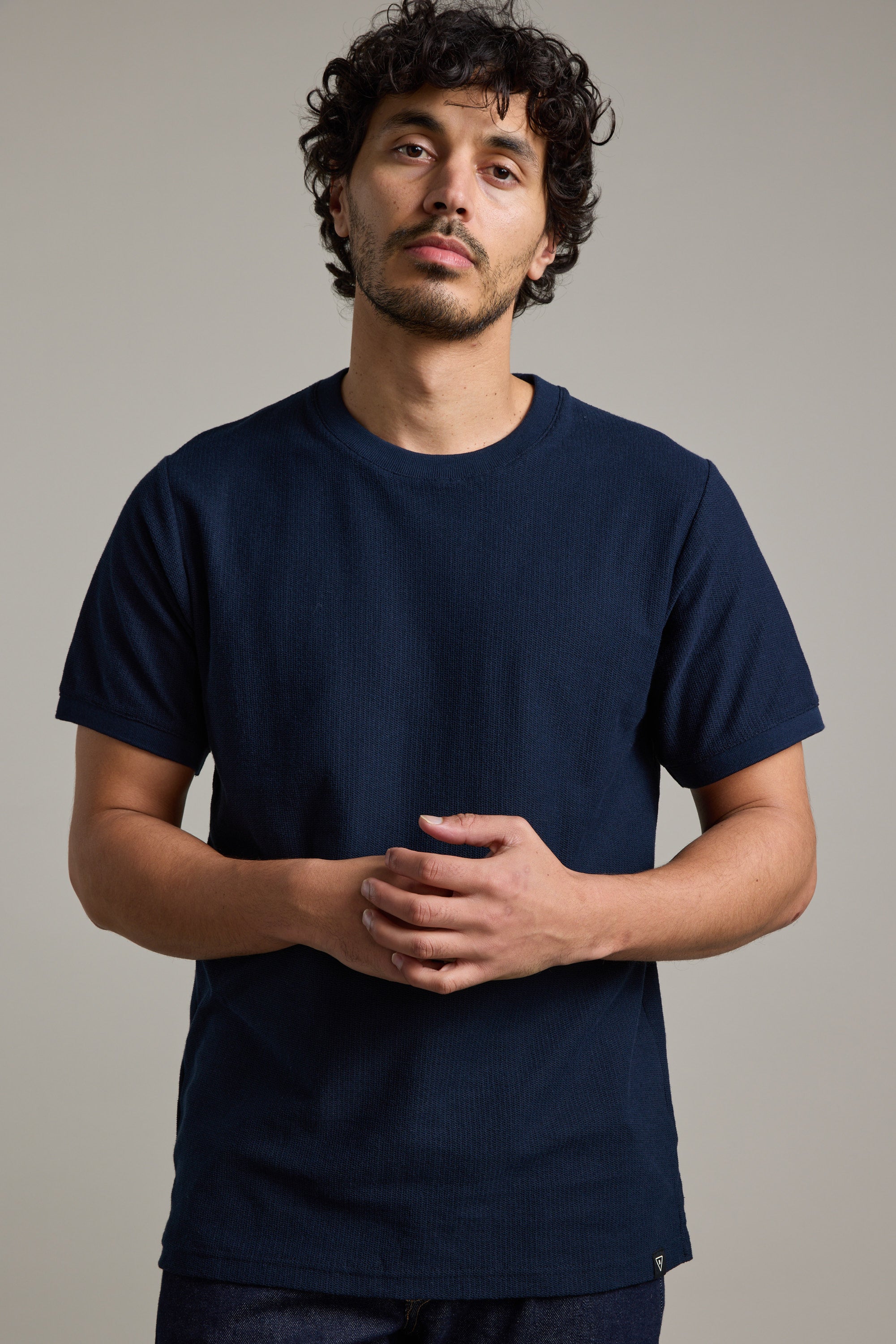 A man with curly dark hair and a mustache wears the Barkers Signature Knit Tee in dark blue, featuring ribbed cuffs. He stands against a plain light background, looking at the camera with his hands clasped in front of him.
