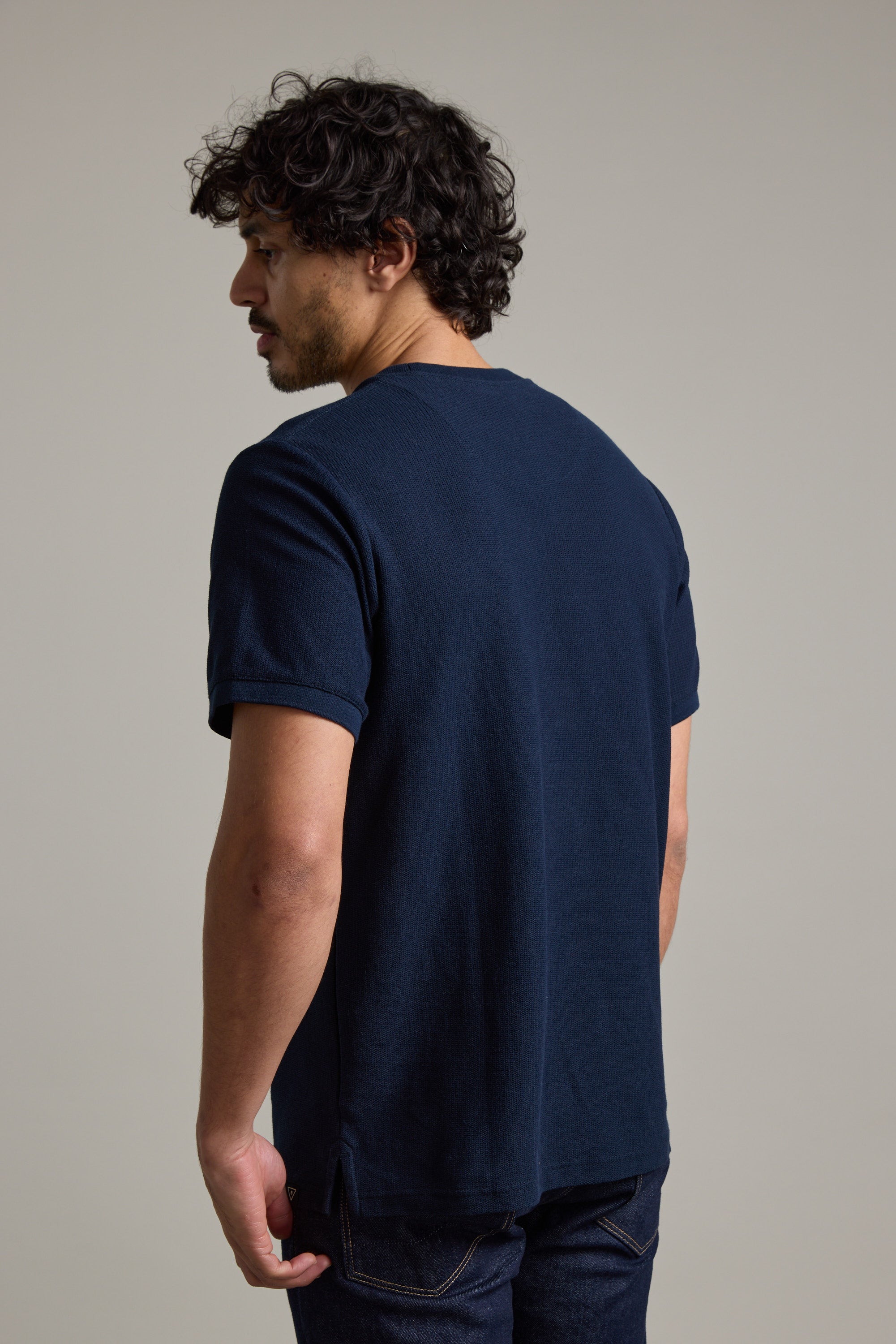 A man with curly dark hair, wearing a navy Barkers Signature Knit Tee with ribbed cuffs and dark jeans, stands against a plain light gray background, looking slightly over his left shoulder.