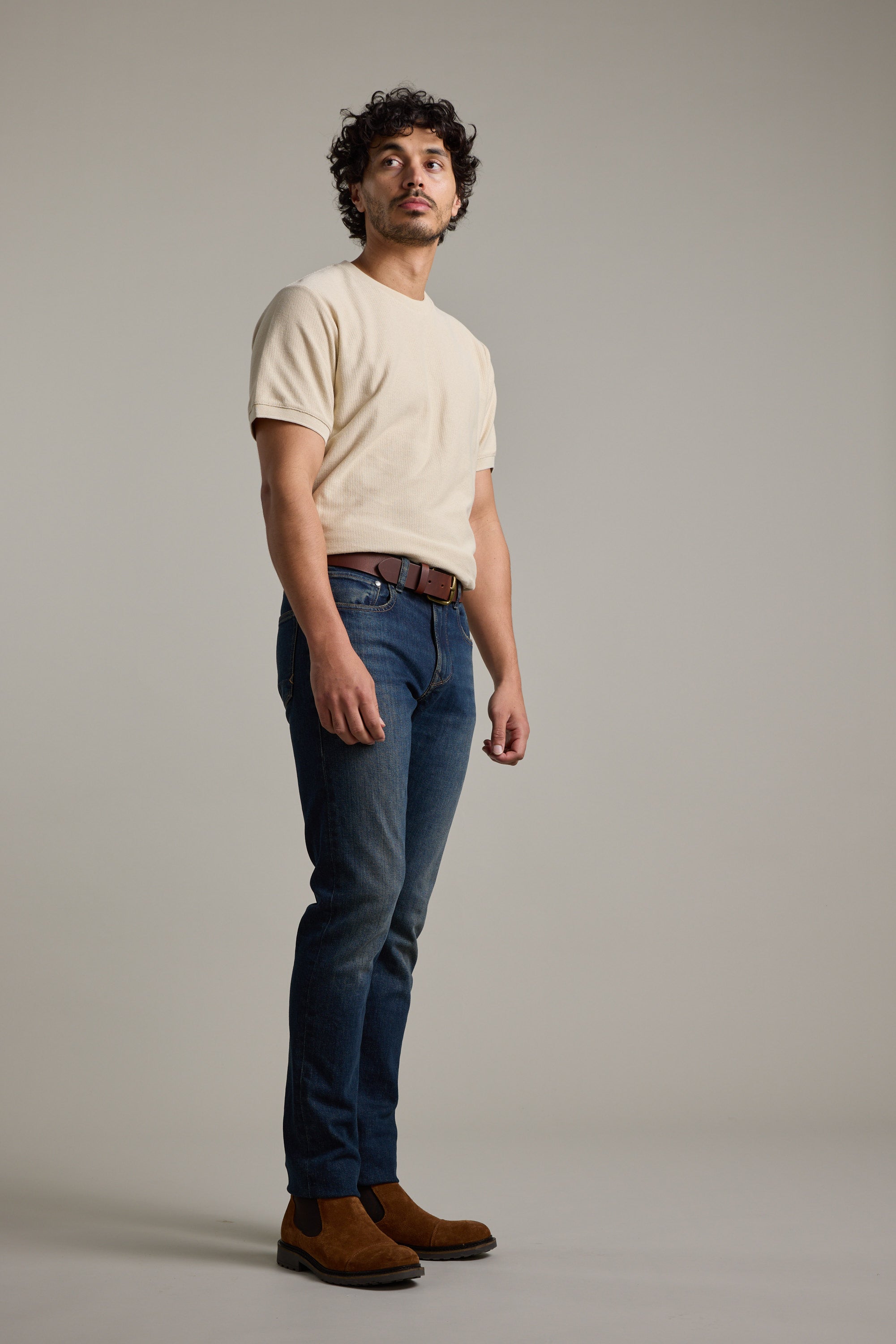 A man with curly hair stands against a plain background wearing Barkers' Signature Knit Tee in beige, blue jeans, a brown belt, and brown suede shoes. He gazes slightly upward with a neutral expression.