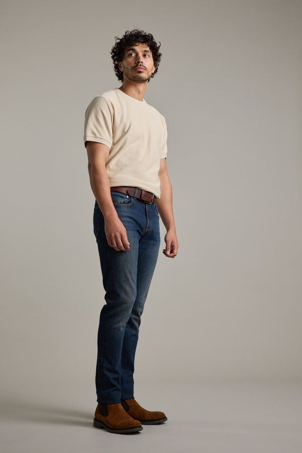 A man with curly hair stands against a plain background wearing Barkers' Signature Knit Tee in beige, blue jeans, a brown belt, and brown suede shoes. He gazes slightly upward with a neutral expression.