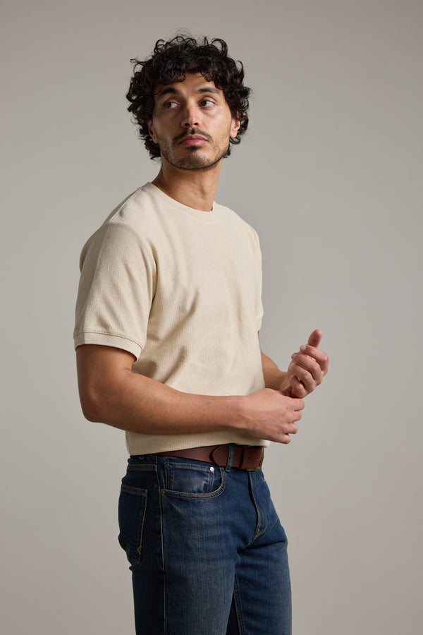 A man with curly dark hair stands against a plain light gray background, wearing a beige Barkers Signature Knit Tee and blue jeans, gazing thoughtfully to his left.