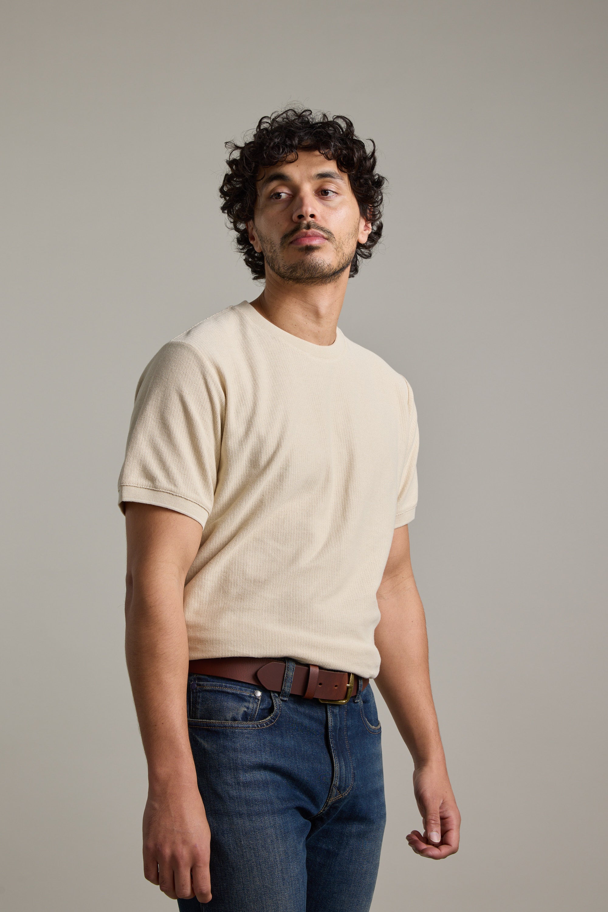 A man with curly dark hair and a short beard wears a Barkers Signature Knit Tee in beige, paired with blue jeans and a brown belt, standing against a plain light gray background and looking slightly to his left.