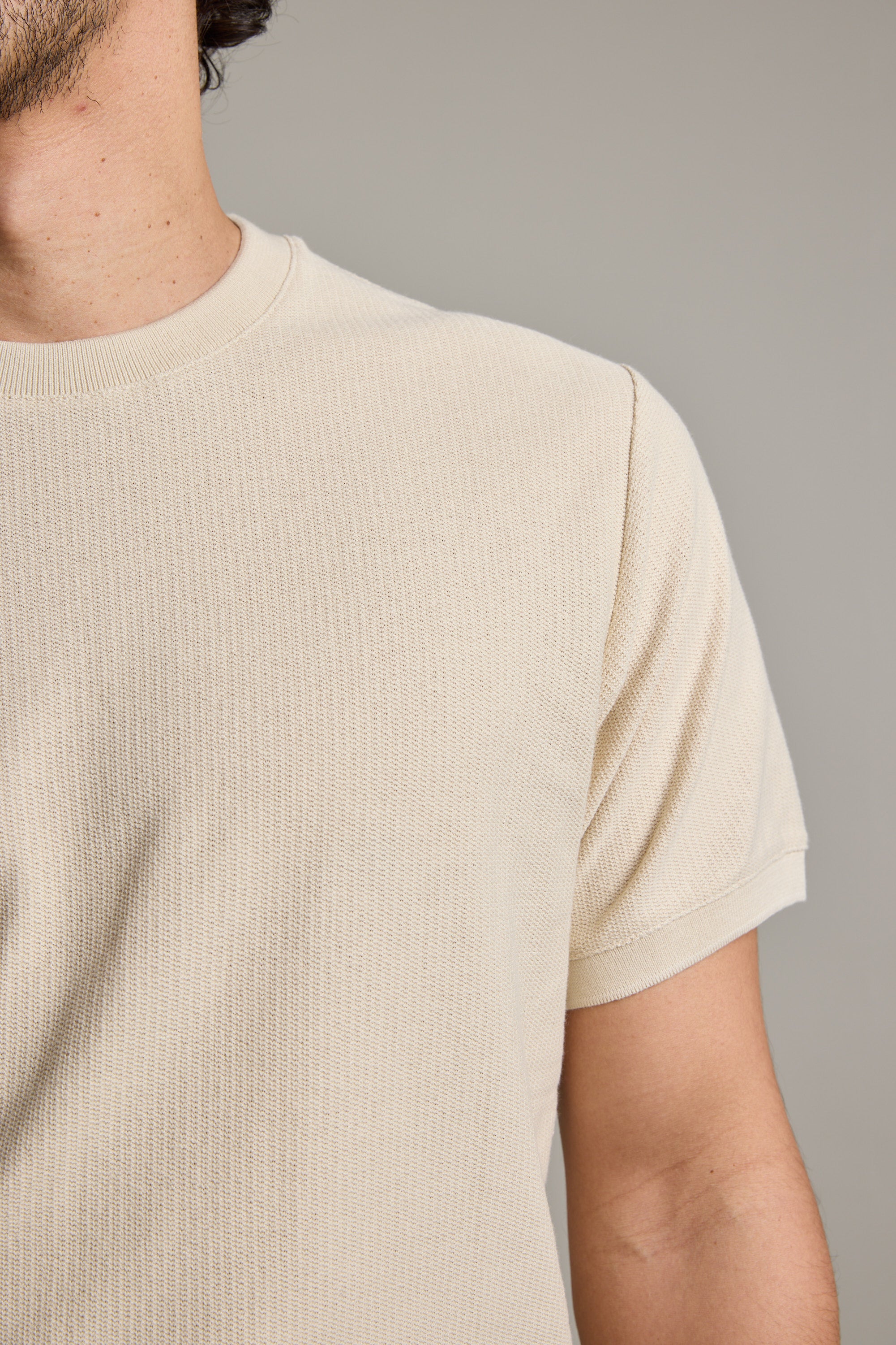 A close-up shows a person wearing Barkers' Signature Knit Tee in light beige. Only their neck, shoulder, and upper arm are visible against a plain, neutral background.