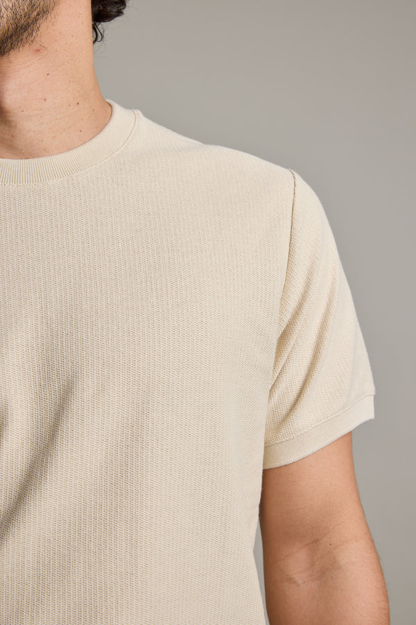 A close-up shows a person wearing Barkers' Signature Knit Tee in light beige. Only their neck, shoulder, and upper arm are visible against a plain, neutral background.