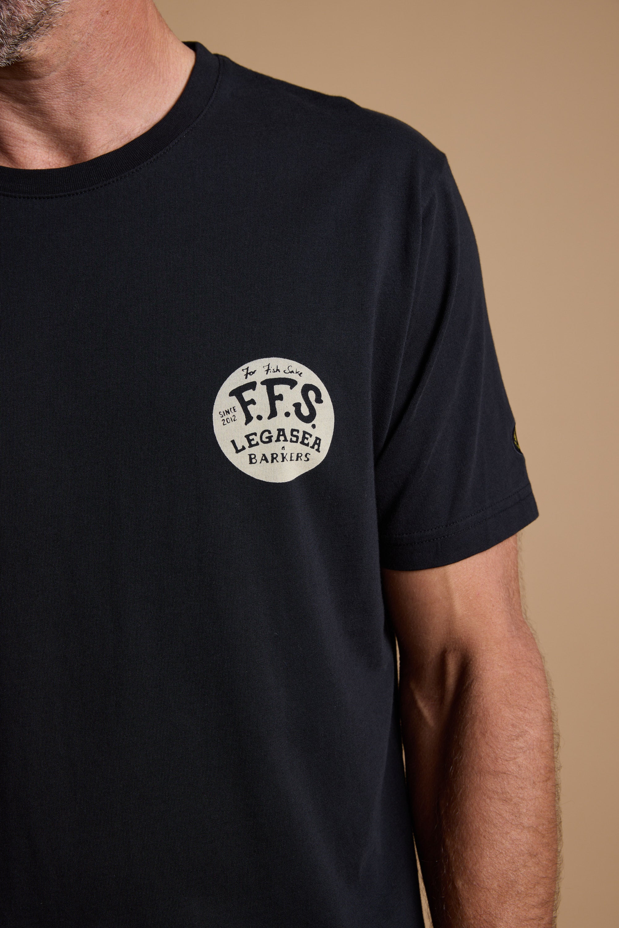 A man in a black Legasea Tail Print Tee, featuring a round white “F.F.S LEGASEA BARKERS” logo and inspired by the New Zealand coastline, stands against a plain beige background with only part of his face and upper body visible.